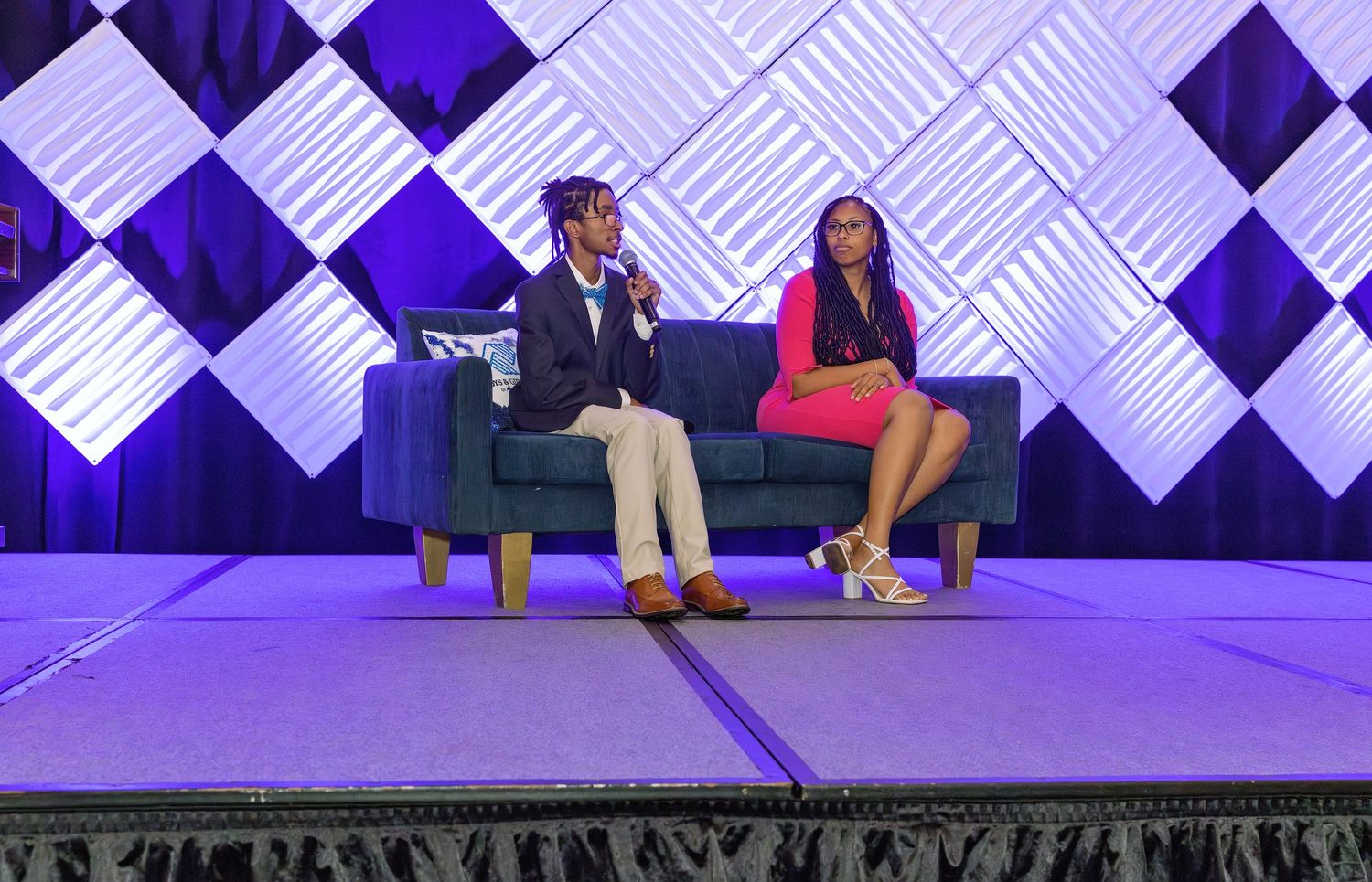 Two people sit on a blue couch on stage with a geometric white and blue backdrop at a conference event.