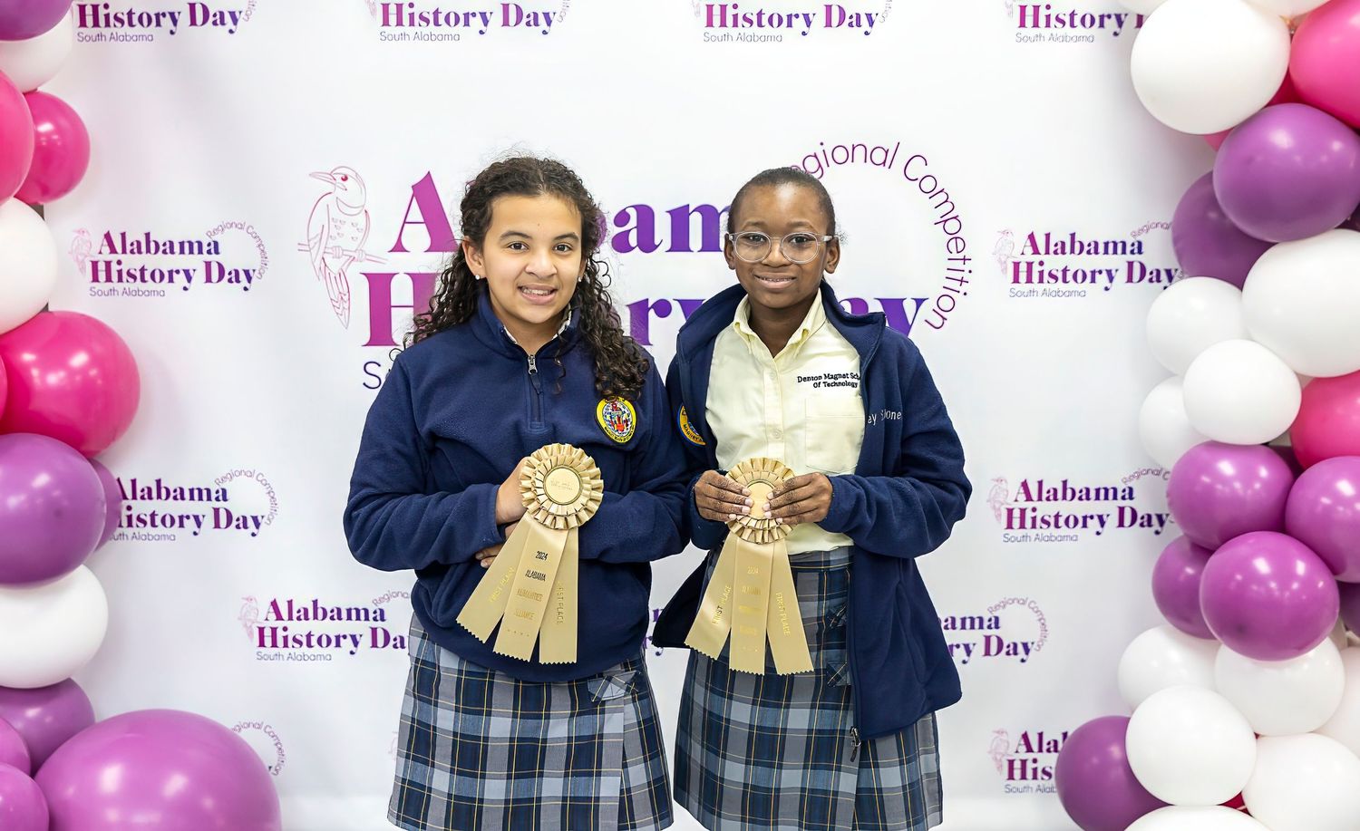Students in navy uniforms display award ribbons at school event with balloon backdrop.