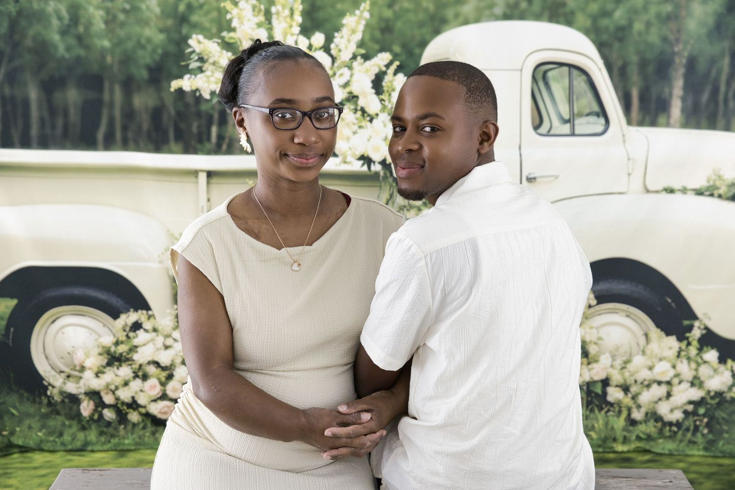 A couple poses together outdoors in front of a vintage white truck and flowering bushes.