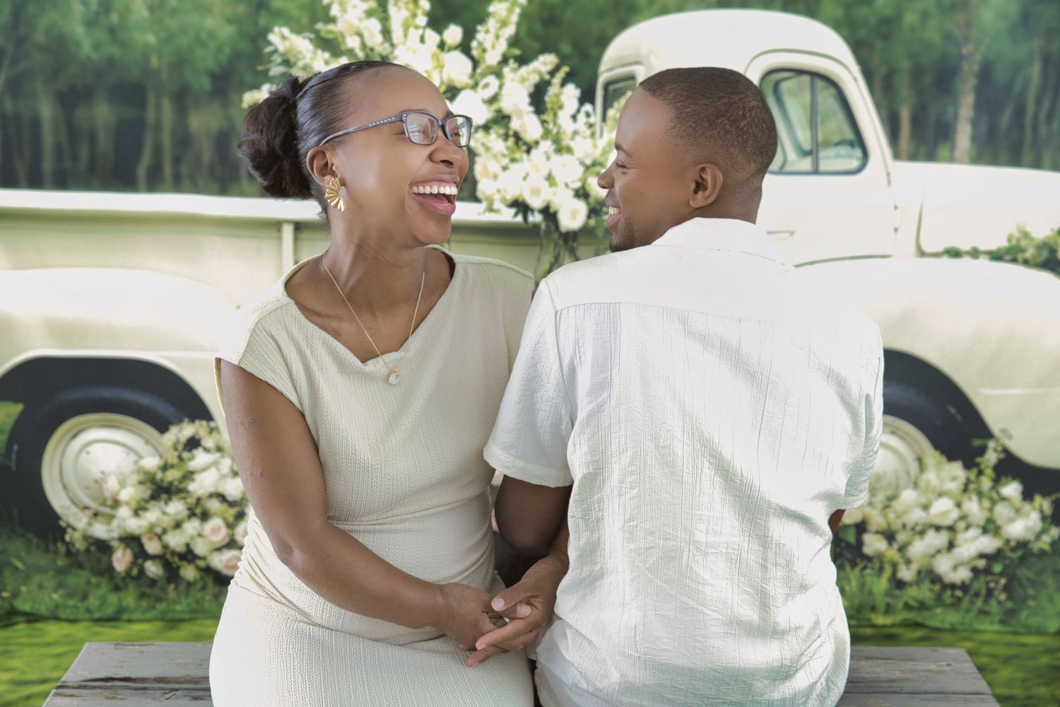 A joyful couple laughs together while sitting outdoors with white flowers and a vintage car in the background.
