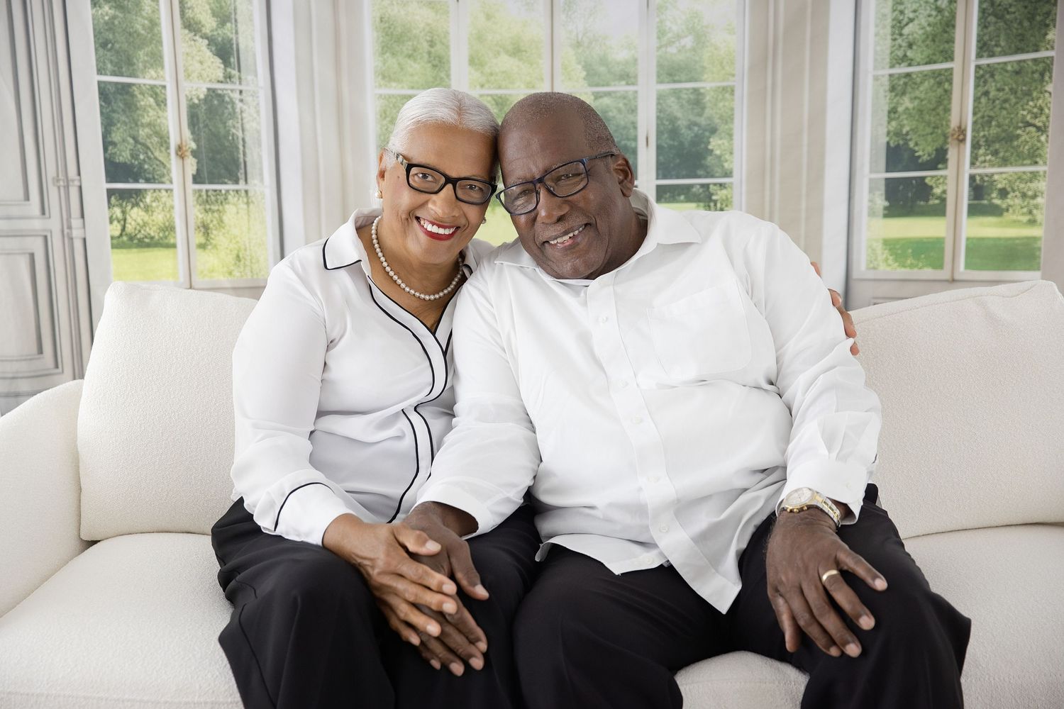 Two people wearing white shirts sit close together on a couch, sharing warm smiles and laughter in a sunlit room.