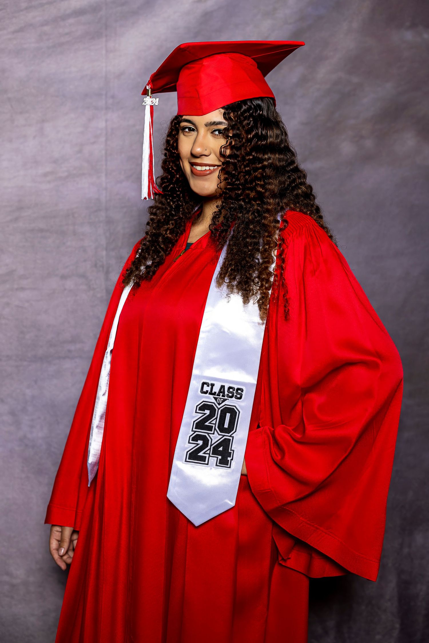 Graduate poses in red cap and gown with white Class of 2024 graduation sash against gray backdrop.