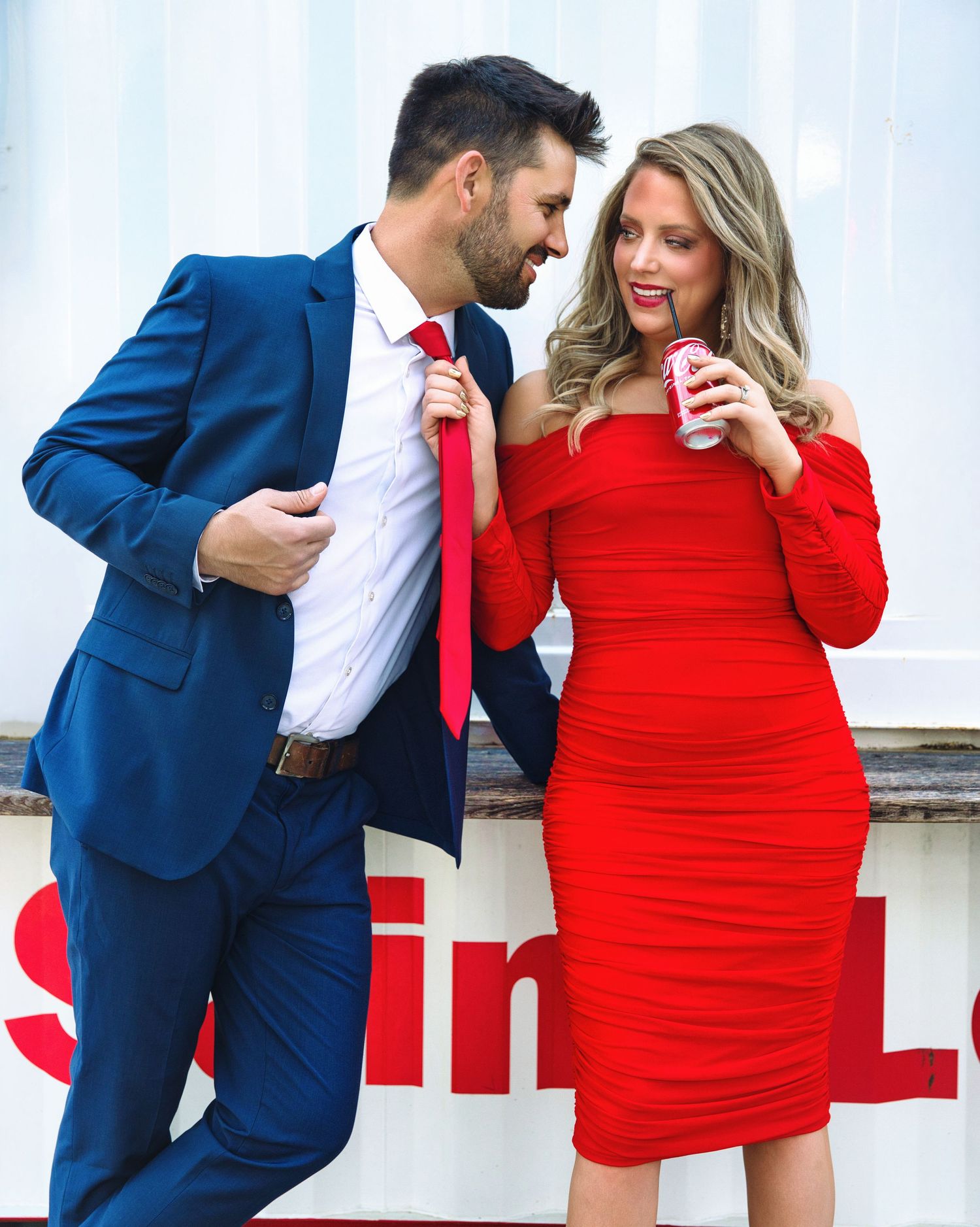 Stylish couple in blue suit and red dress share playful moment while holding Coca-Cola.