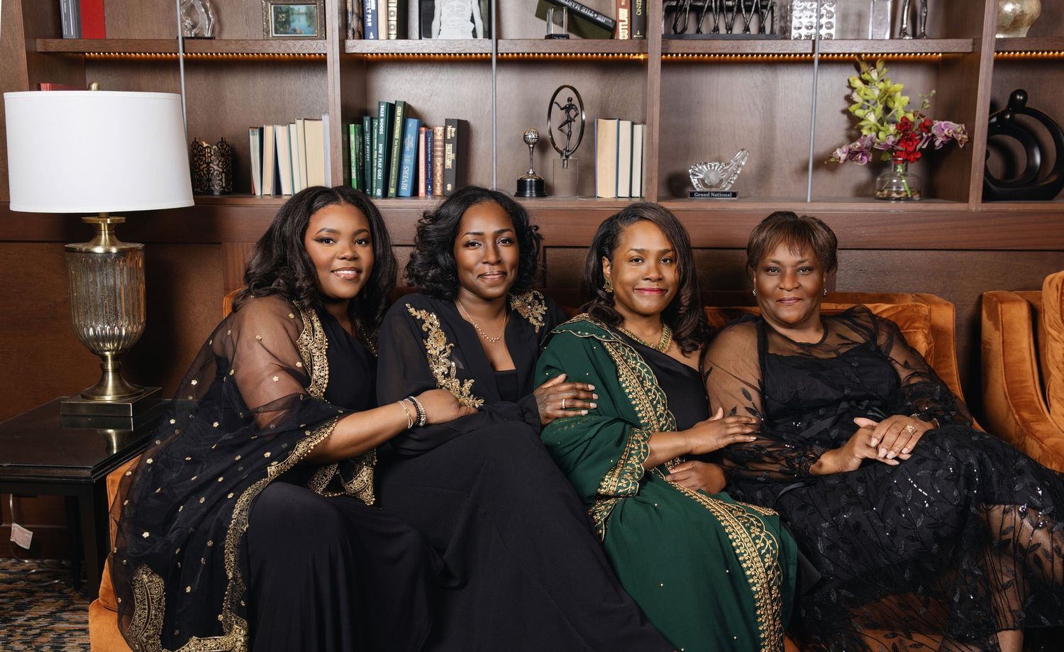 A group of four people wearing elegant evening dresses pose together on a couch in a cozy living room setting.