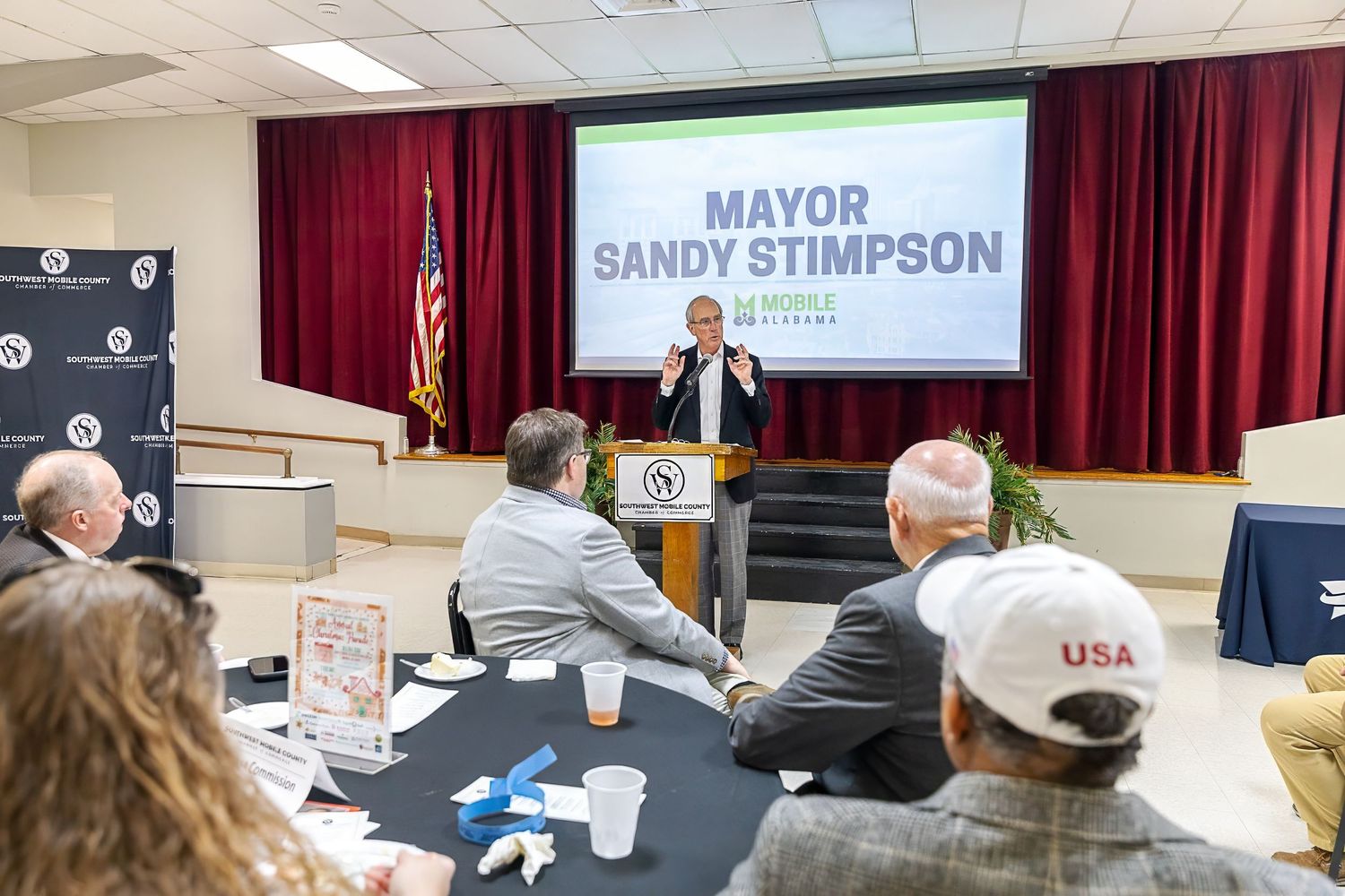 A speaker stands at a podium during a political event with 'Mayor Sandy's Simpson' signage and attendees seated at tables.