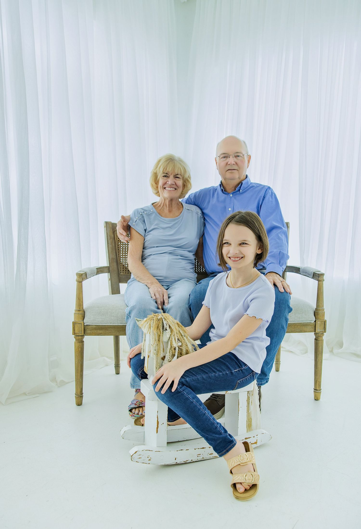Three generations pose for a family portrait, two grandparents and a young girl, in a bright white studio setting.