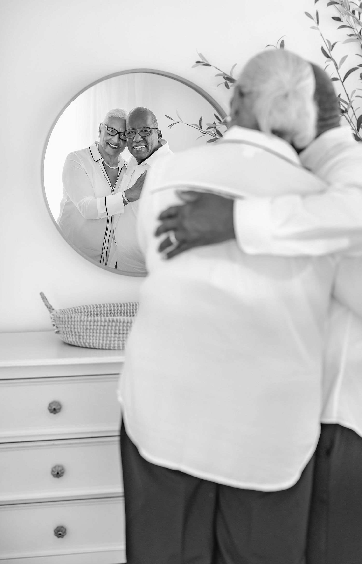 A tender black and white reflection in a mirror shows a couple embracing while standing next to a white dresser.