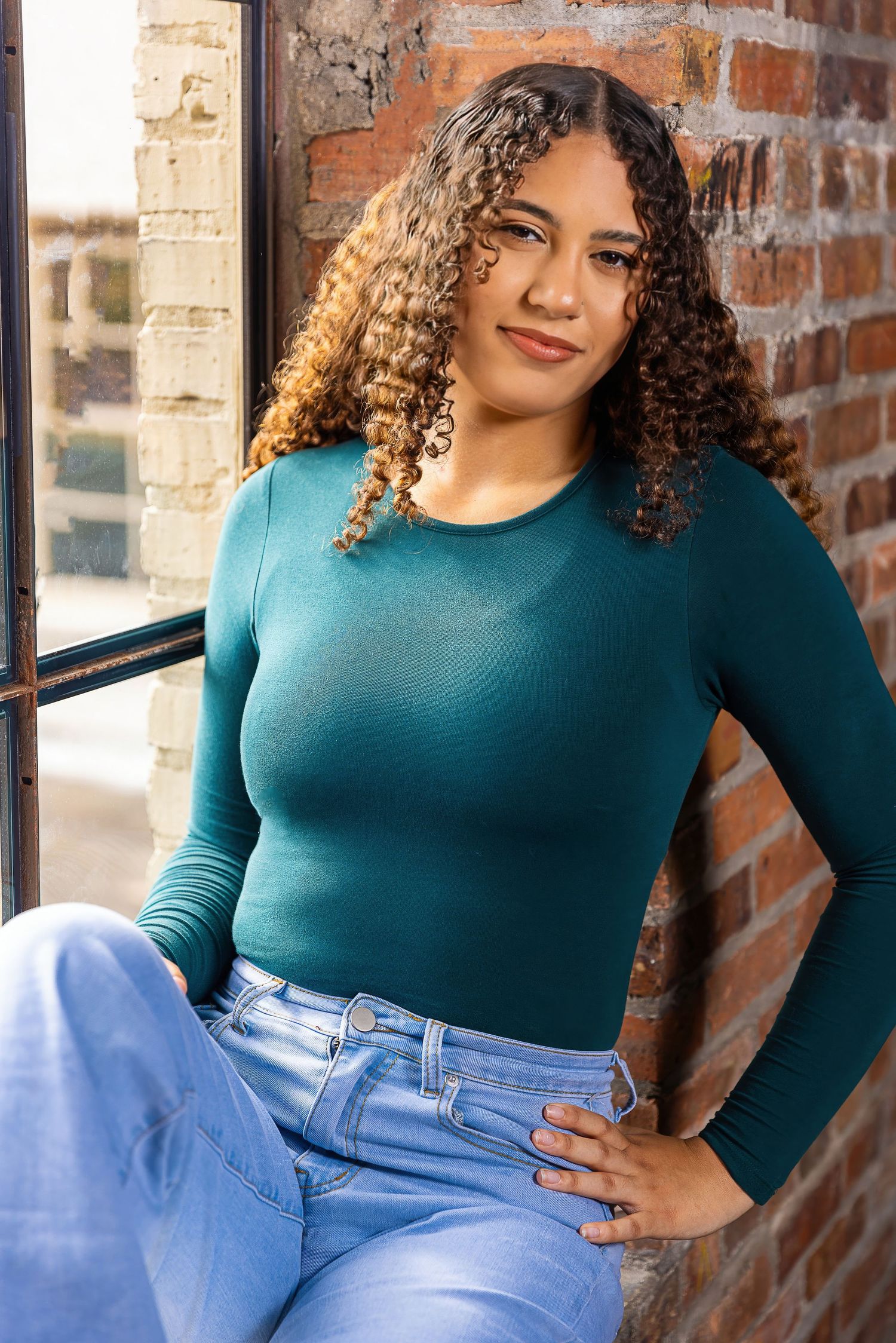 Woman in teal long sleeve top and blue jeans poses against brick wall in natural lighting.