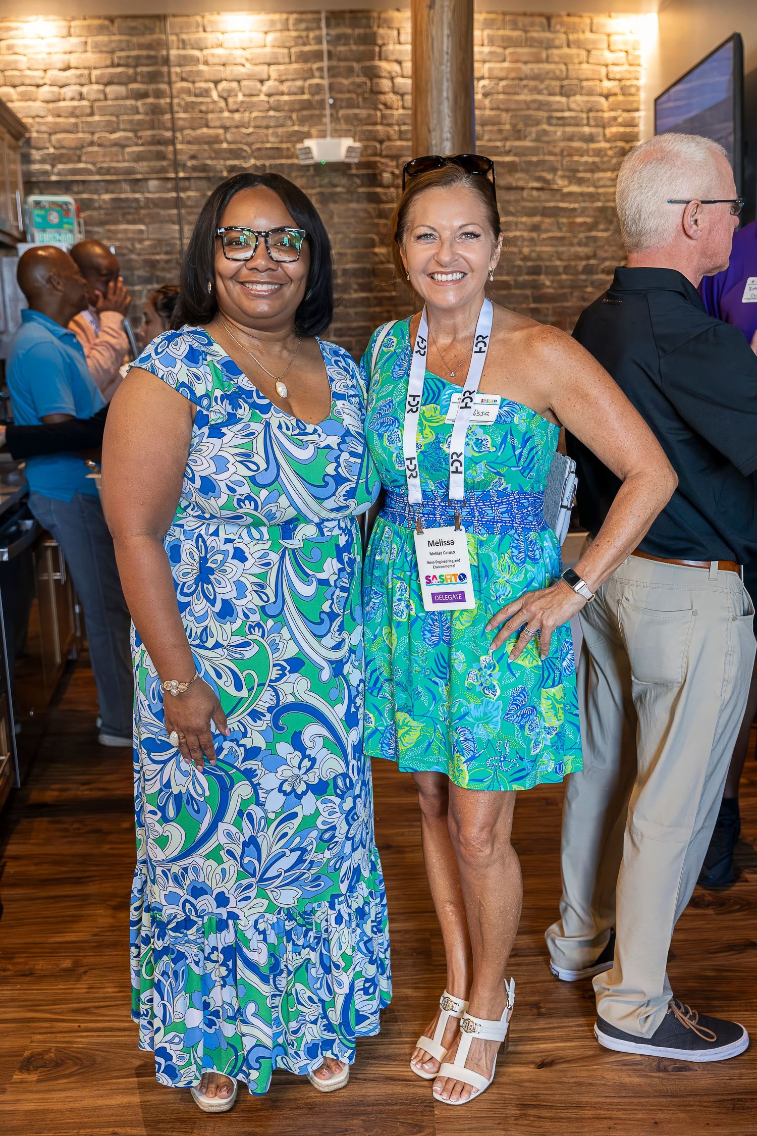 Two women in colorful floral dresses posing together at social gathering.
