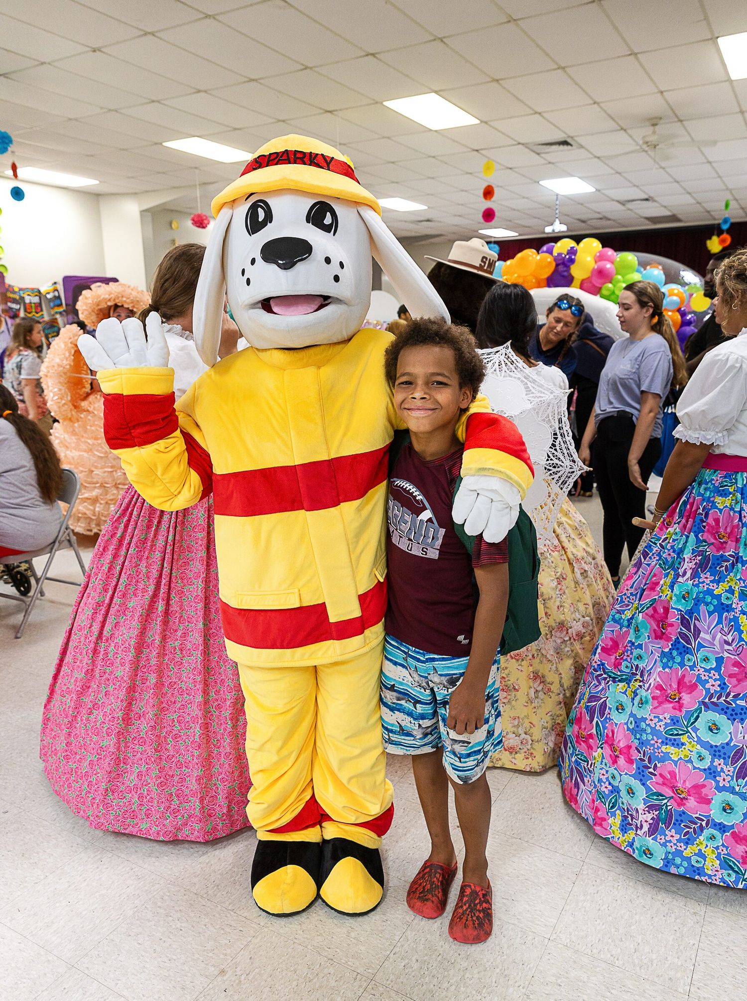 Mascot character in yellow costume poses with child at festive indoor event.