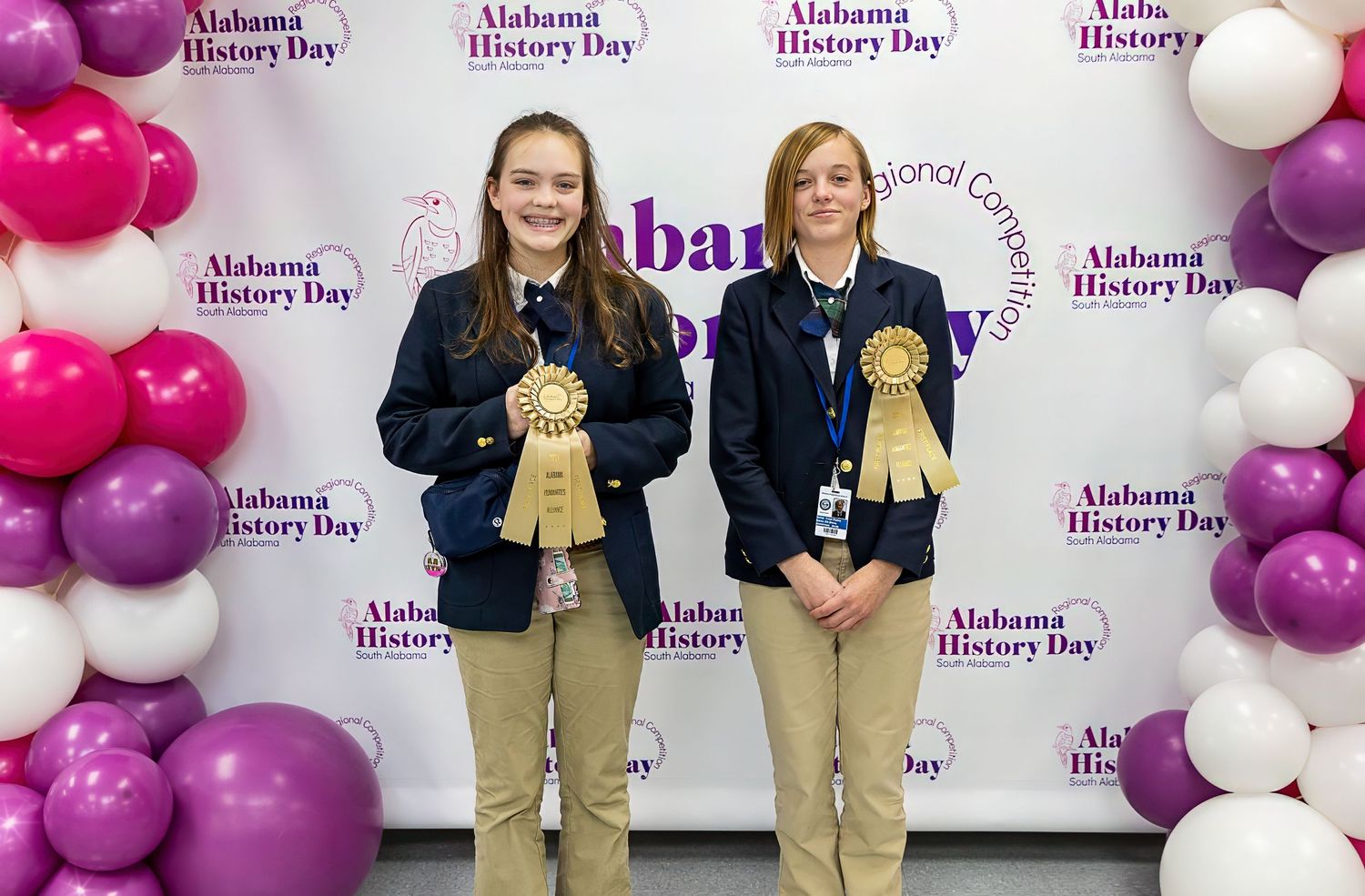 Two students holding award ribbons in front of pink and purple balloon display.
