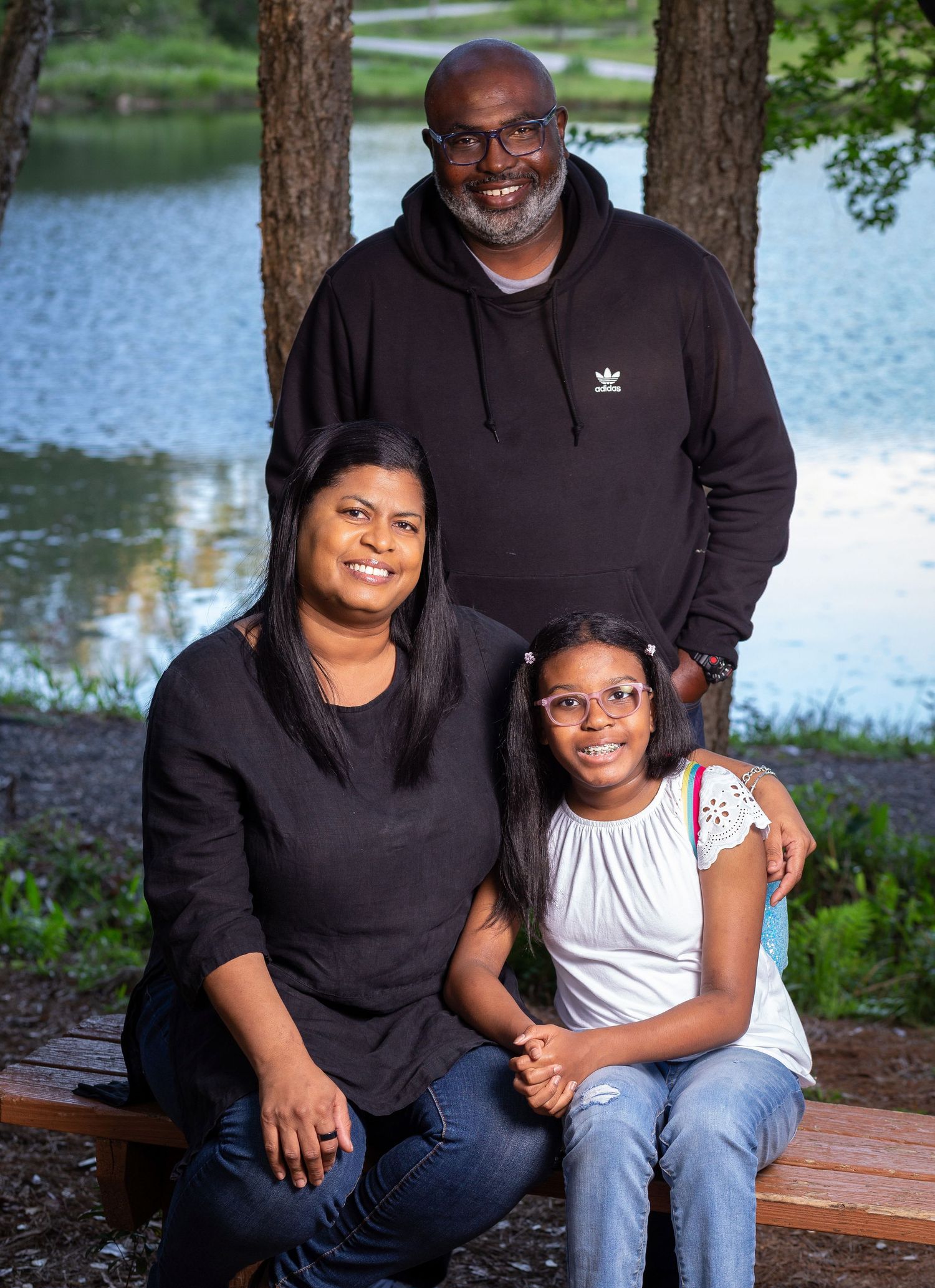 Family portrait of three people sitting on bench by lake wearing dark hoodies and casual attire.