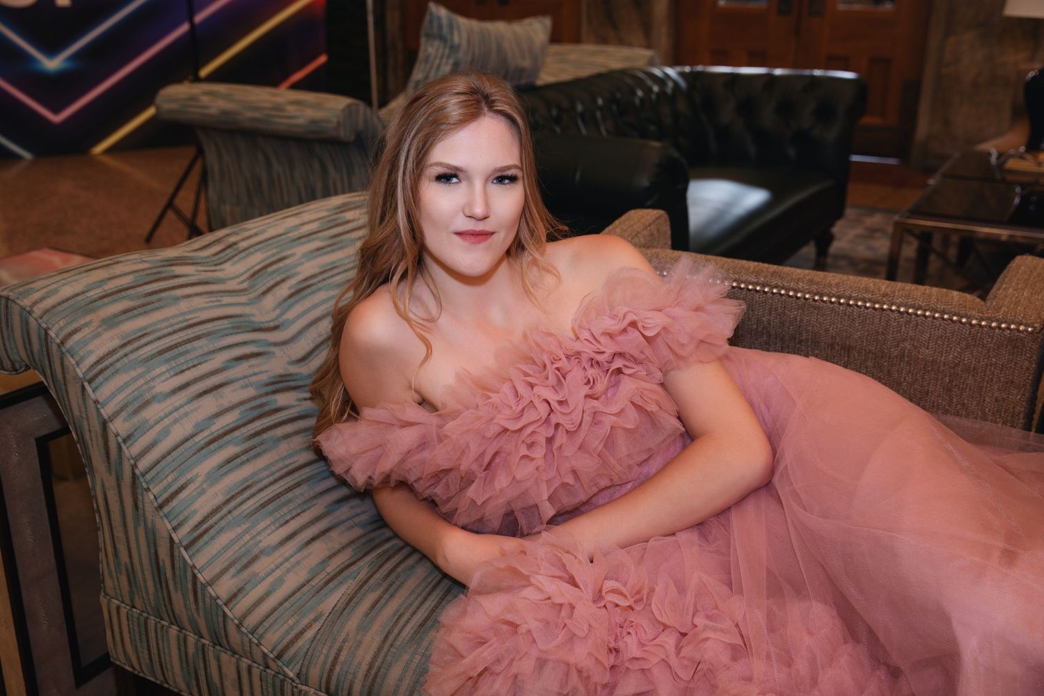 A portrait of someone wearing a flowing pink tulle gown while reclining on a striped couch.