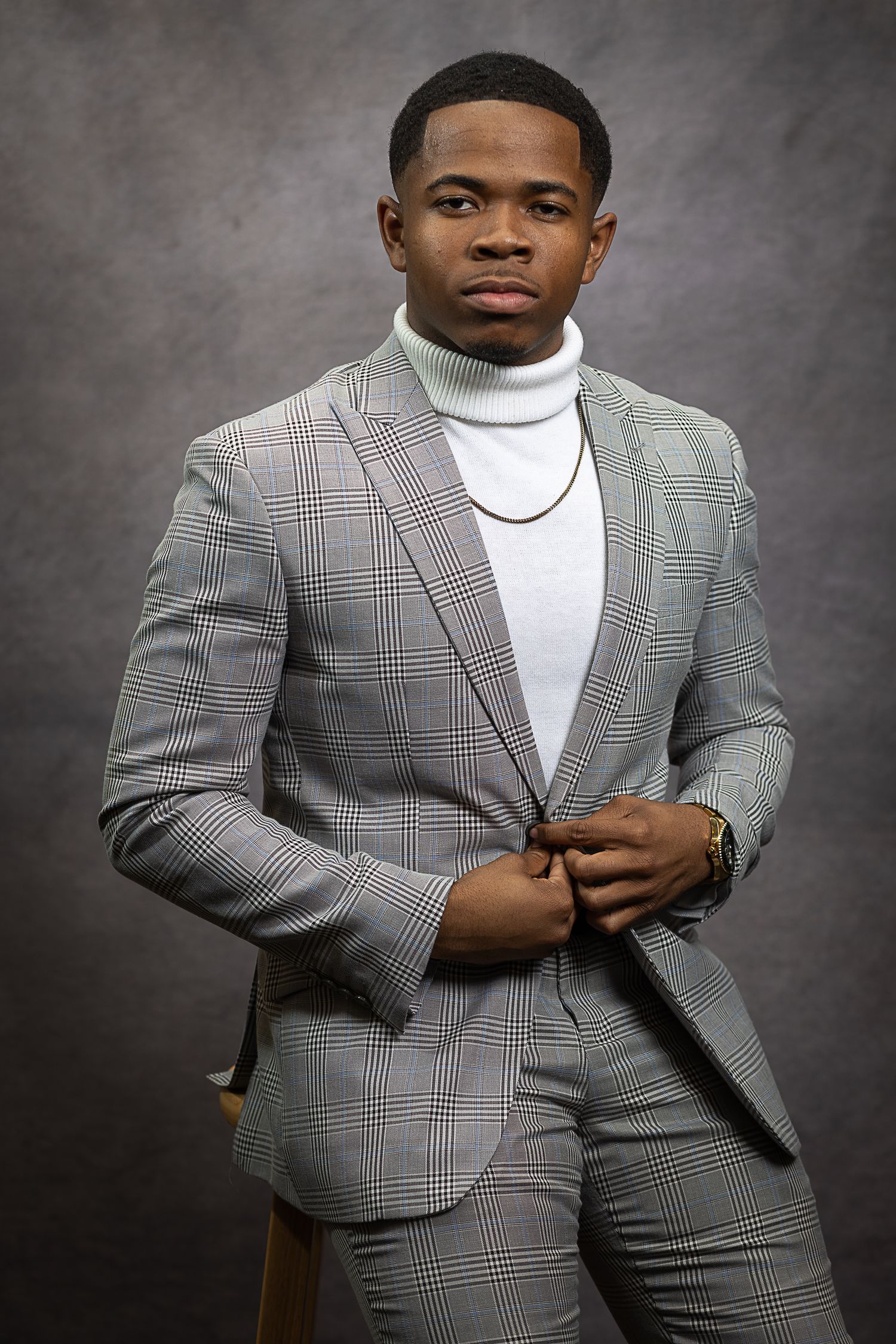 Professional portrait of person in plaid grey suit with white turtleneck against dark backdrop.
