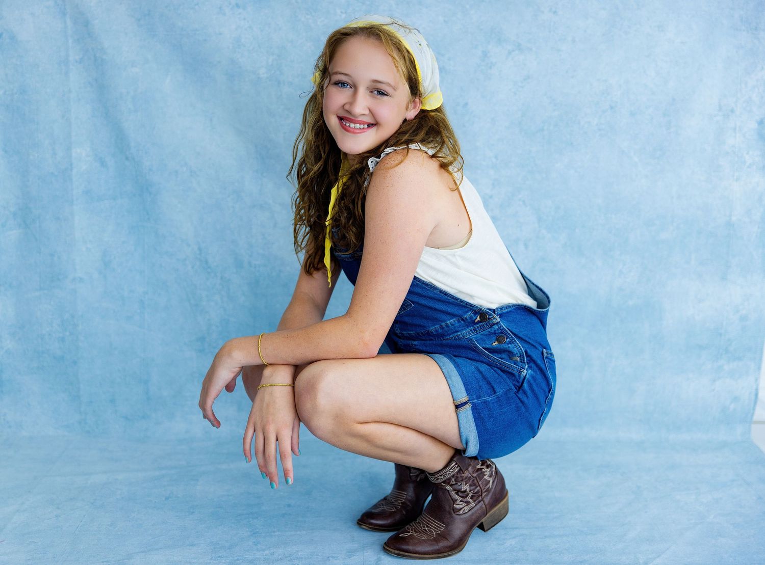 Young girl in denim overalls and boots crouching and smiling against a blue studio backdrop.