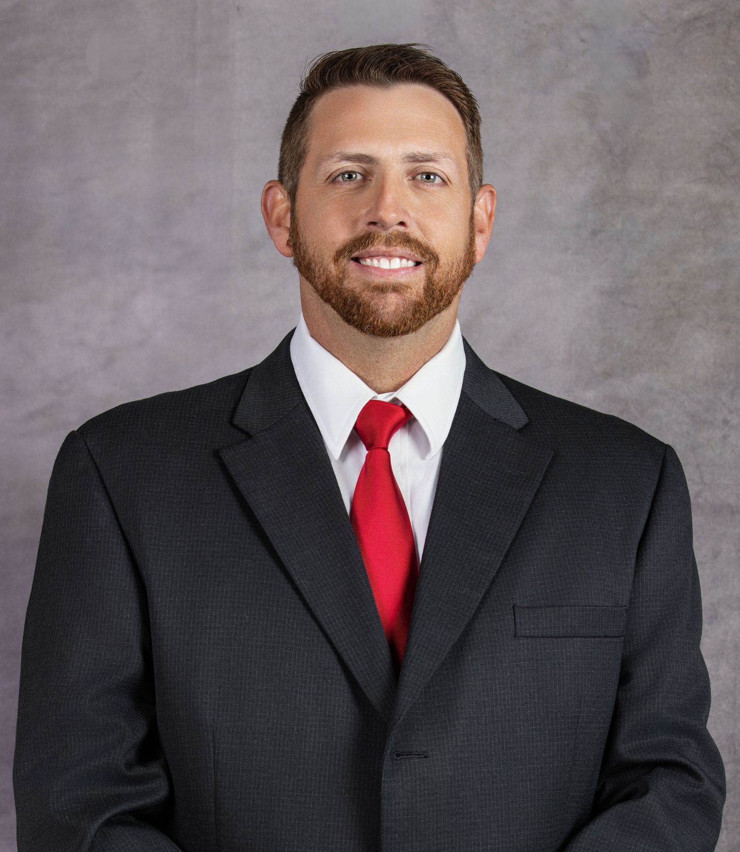Professional headshot portrait of someone wearing a dark suit and red tie against a gray background.