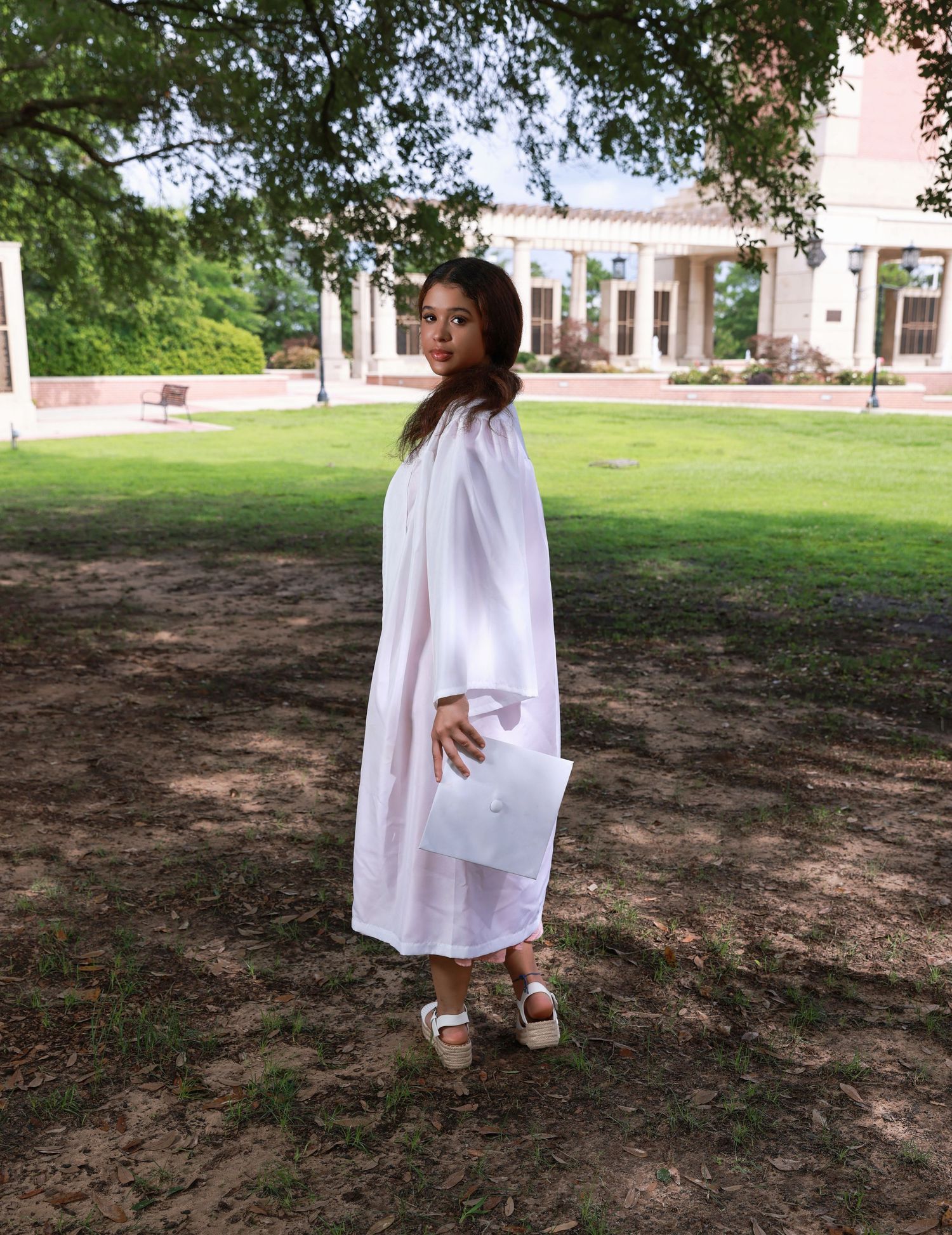 Graduate in white graduation gown stands on lawn in front of columned building during outdoor ceremony.