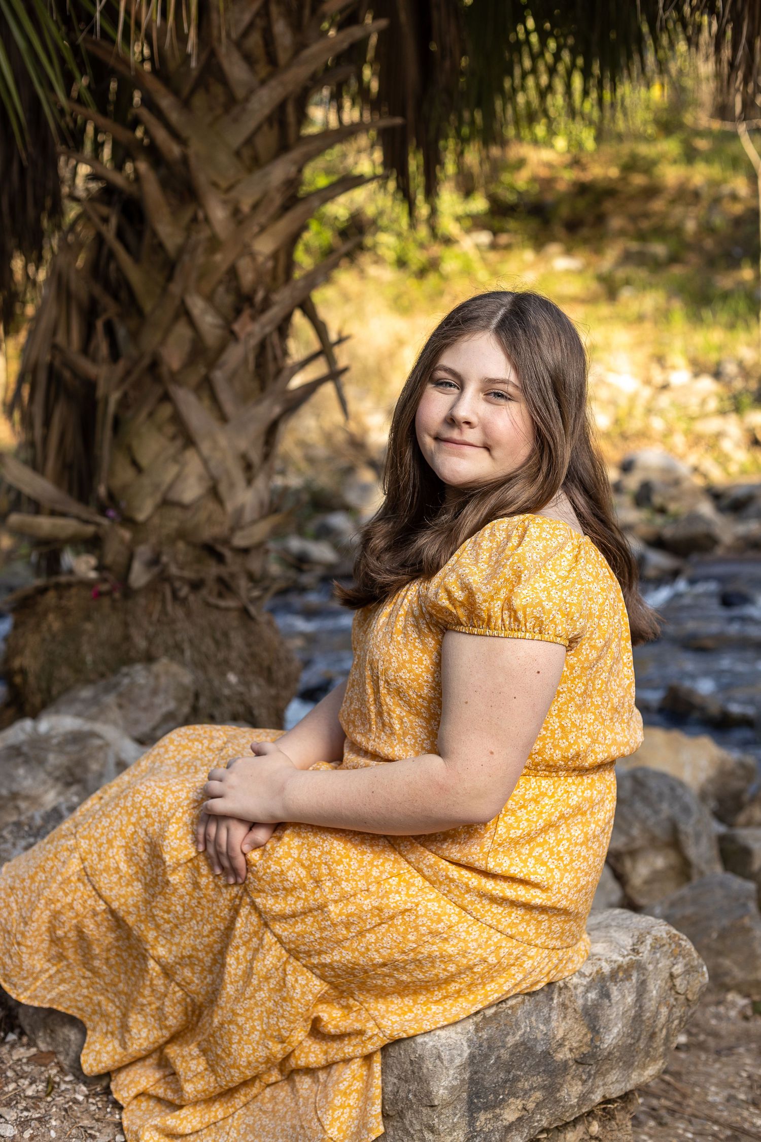 Portrait of a person in a yellow dress sitting on rocks by a creek.