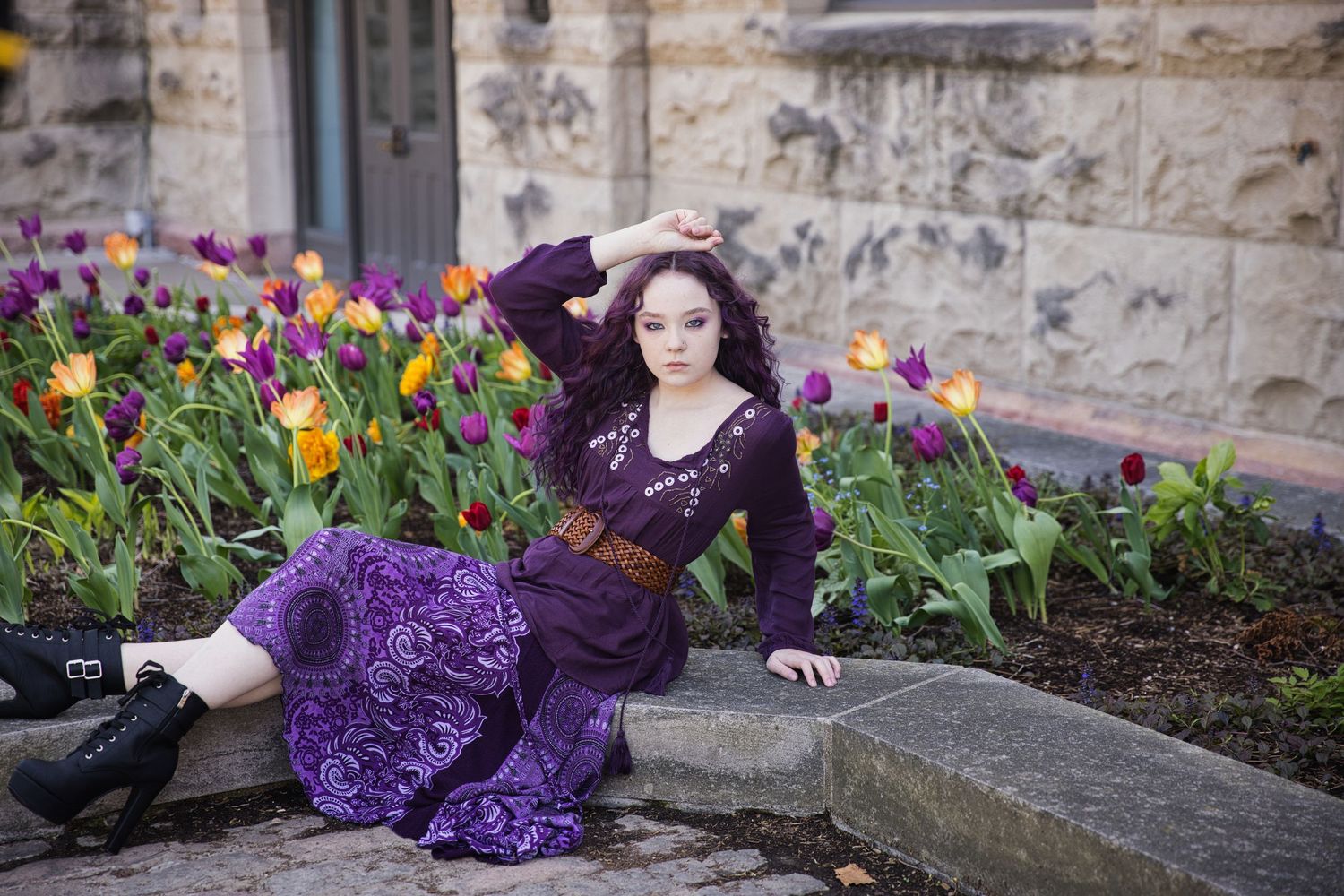 A woman in purple dress reclines among colorful tulips near a stone building.