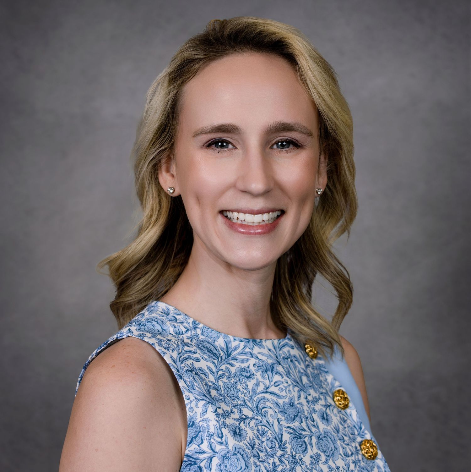 Professional headshot of a person wearing a blue floral sleeveless top against a gray background.