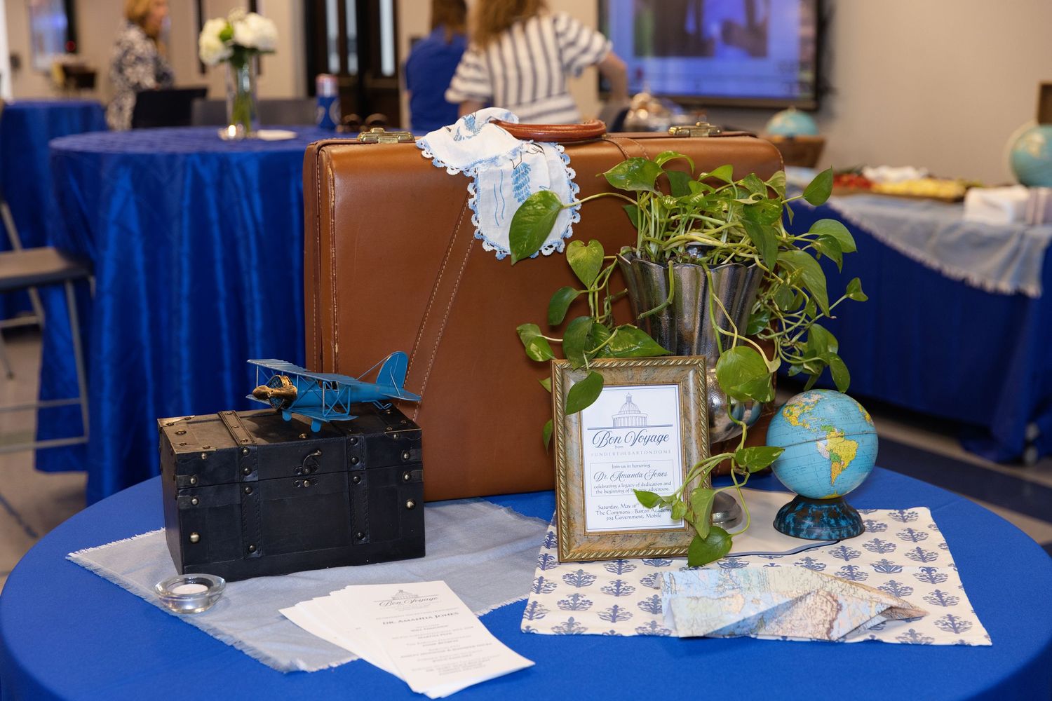 Travel-themed baby shower table with vintage suitcase, toy airplane, globe, and lush greenery on blue tablecloth.