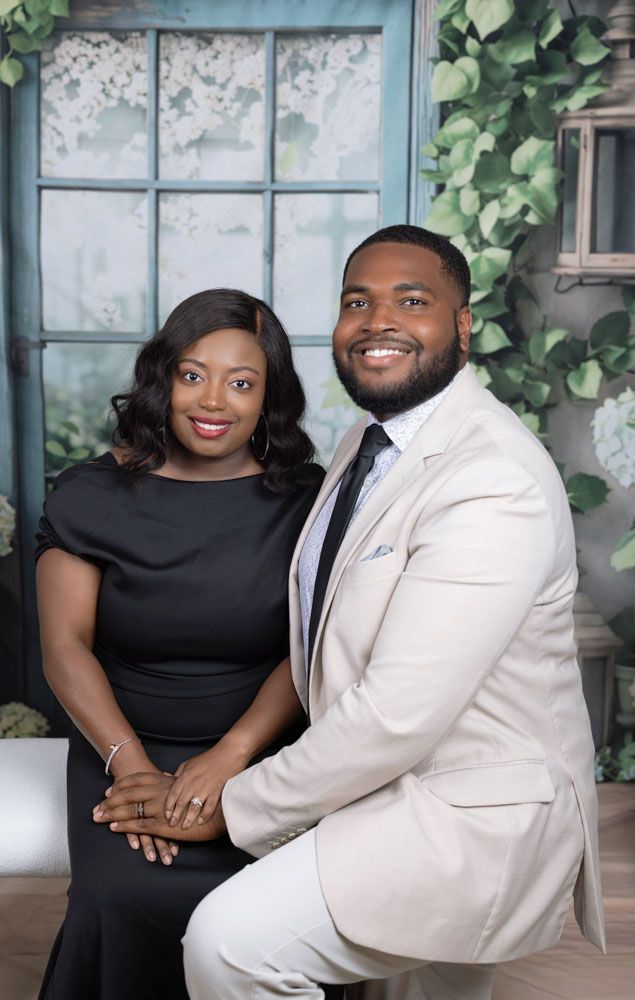 Couple in formal attire pose intimately by window with climbing vines and soft lighting.