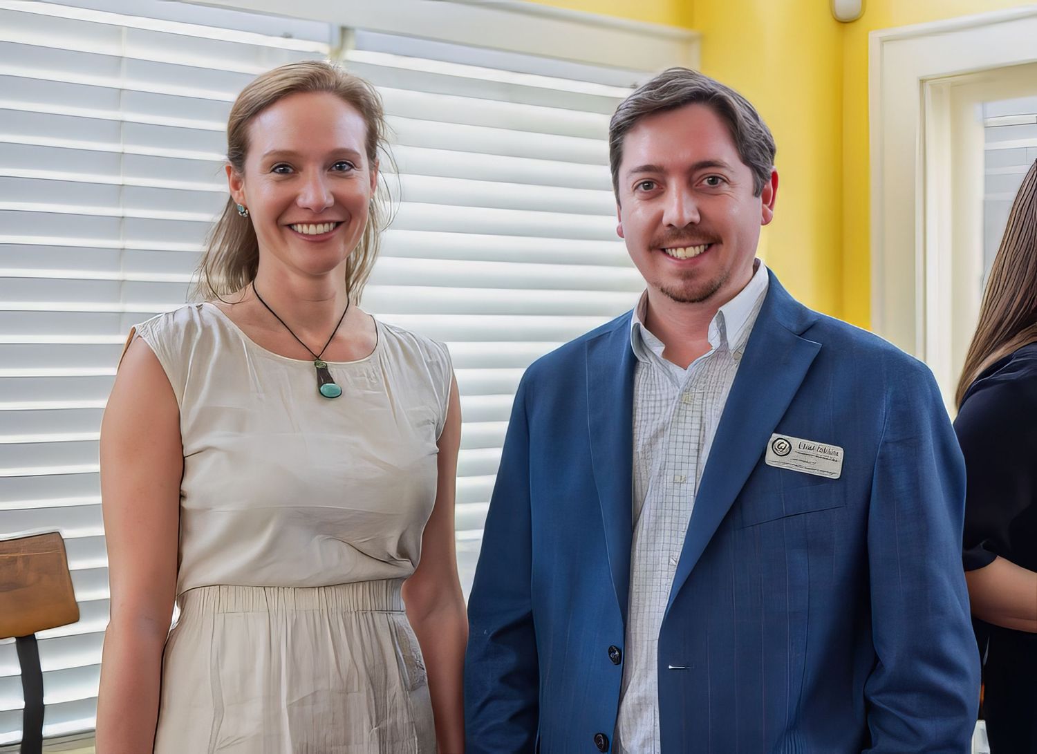 Two professionally dressed people smile while standing together in front of a yellow wall with white window blinds.