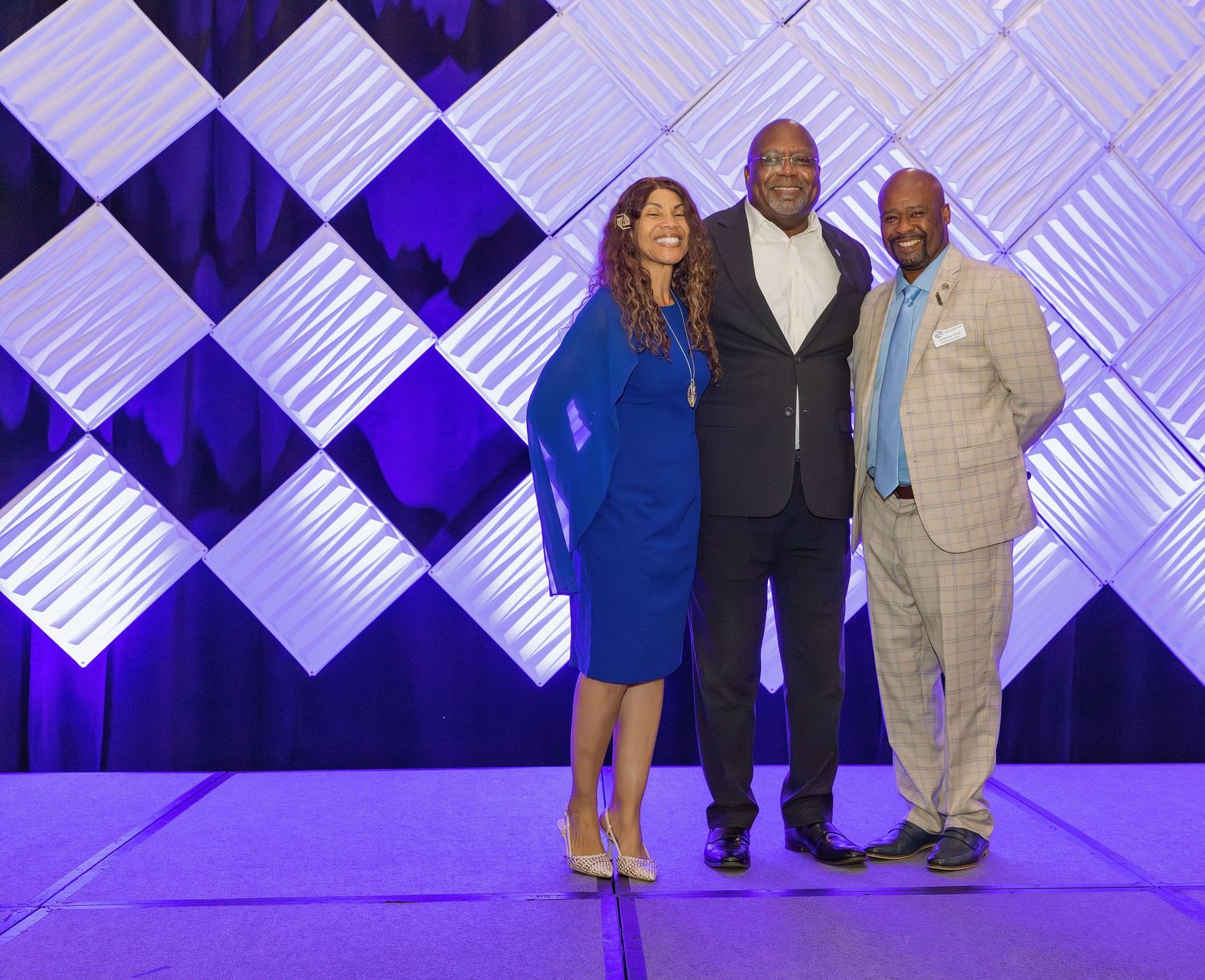 Three professionals pose together on stage in front of a decorative geometric white and purple backdrop at a formal event.