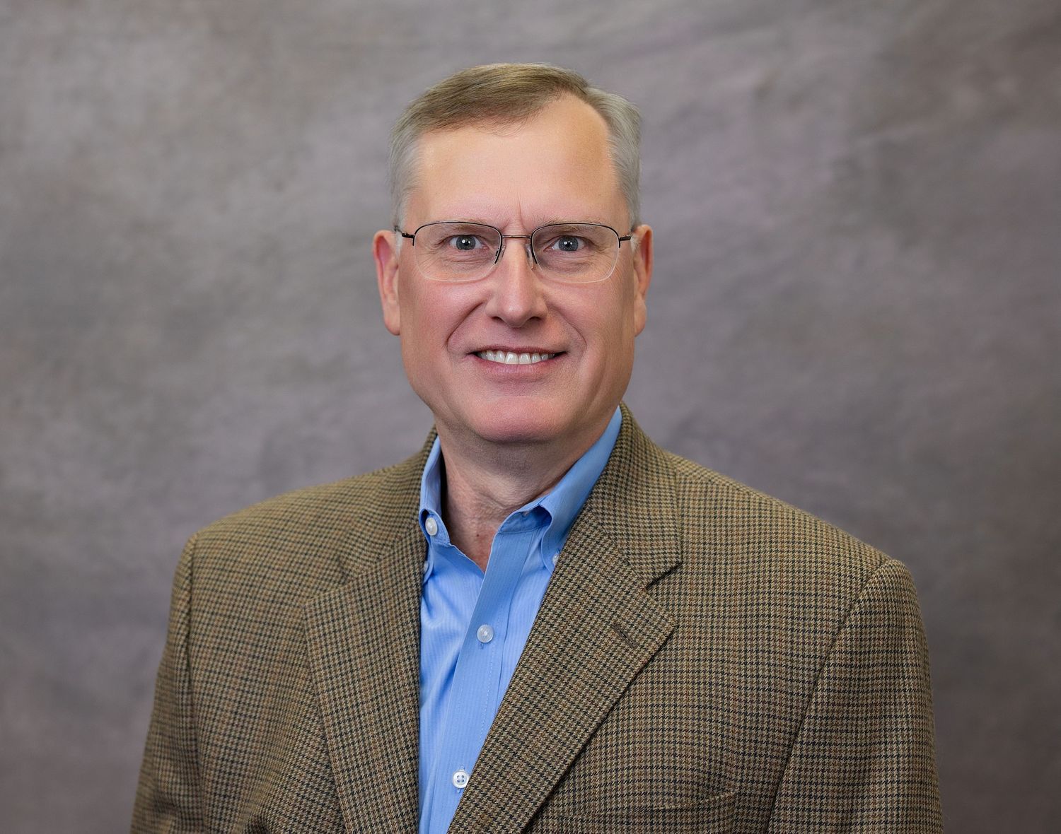 Professional headshot of person wearing glasses, blue dress shirt and brown tweed blazer against grey background.