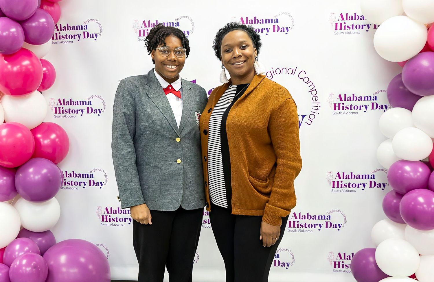 Two individuals pose in professional attire against balloon-decorated backdrop at school event.