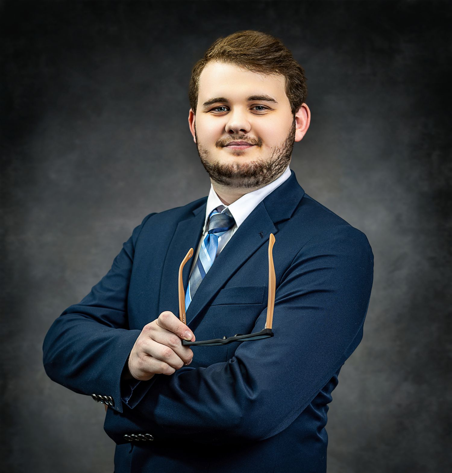Corporate portrait of person in blue suit and tie holding glasses with confident smile.