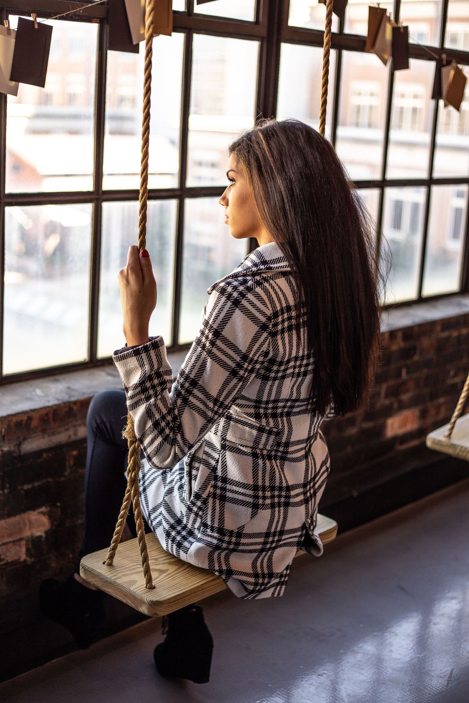 Person in plaid shirt sitting on rope swing by industrial window.