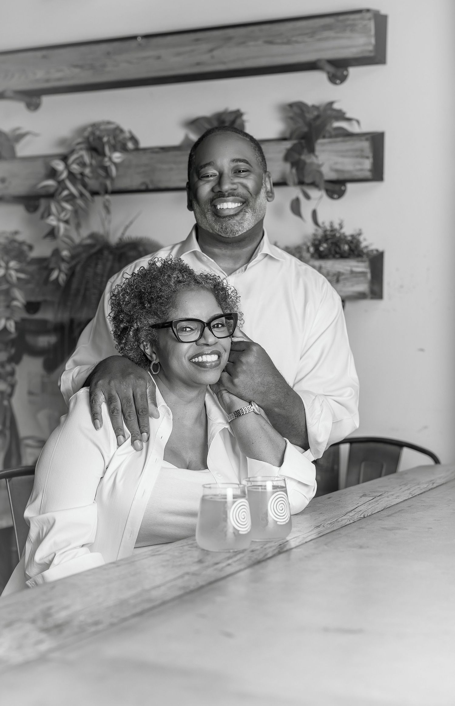 Black and white photo of a smiling couple posing together at a table with hanging plants in the background.