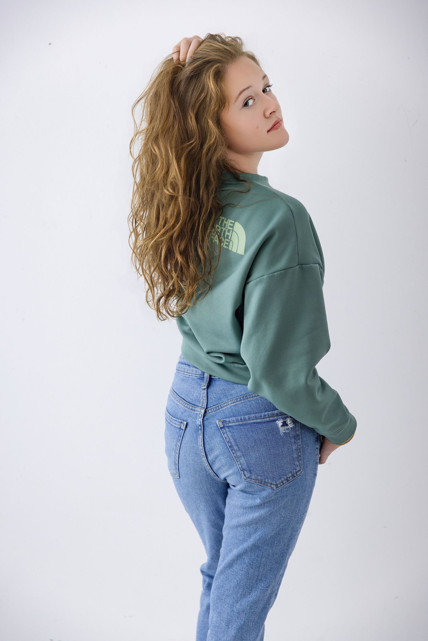 Young woman with curly hair wearing a green North Face sweatshirt and blue jeans, posing against a white background.