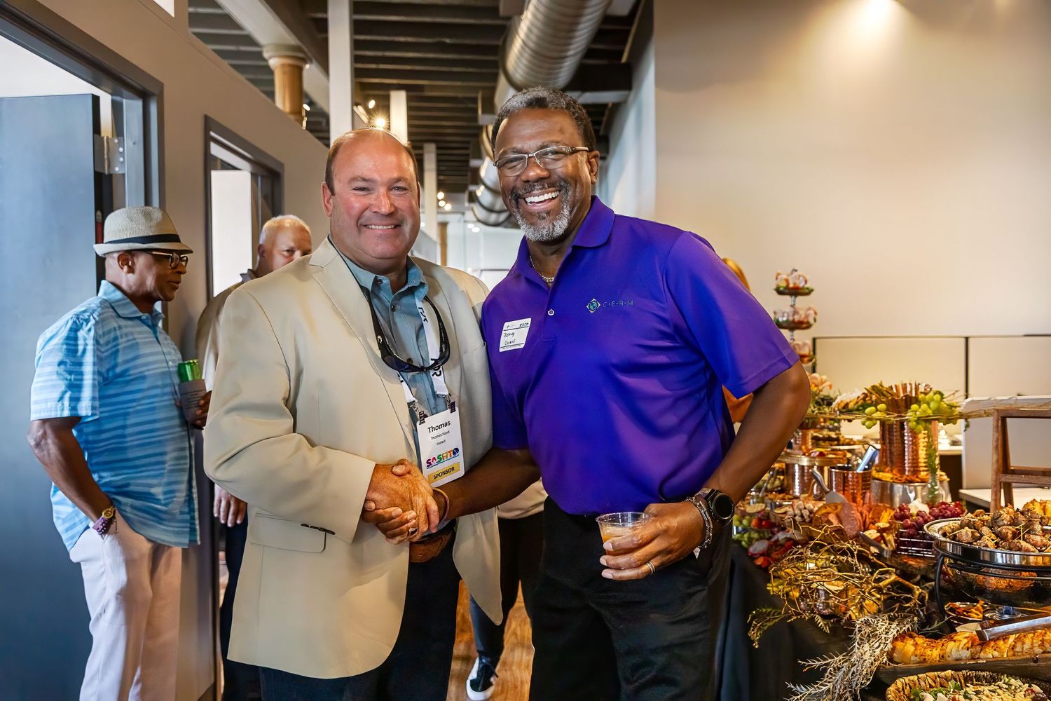 Two people shake hands at casual networking event with buffet table in background.