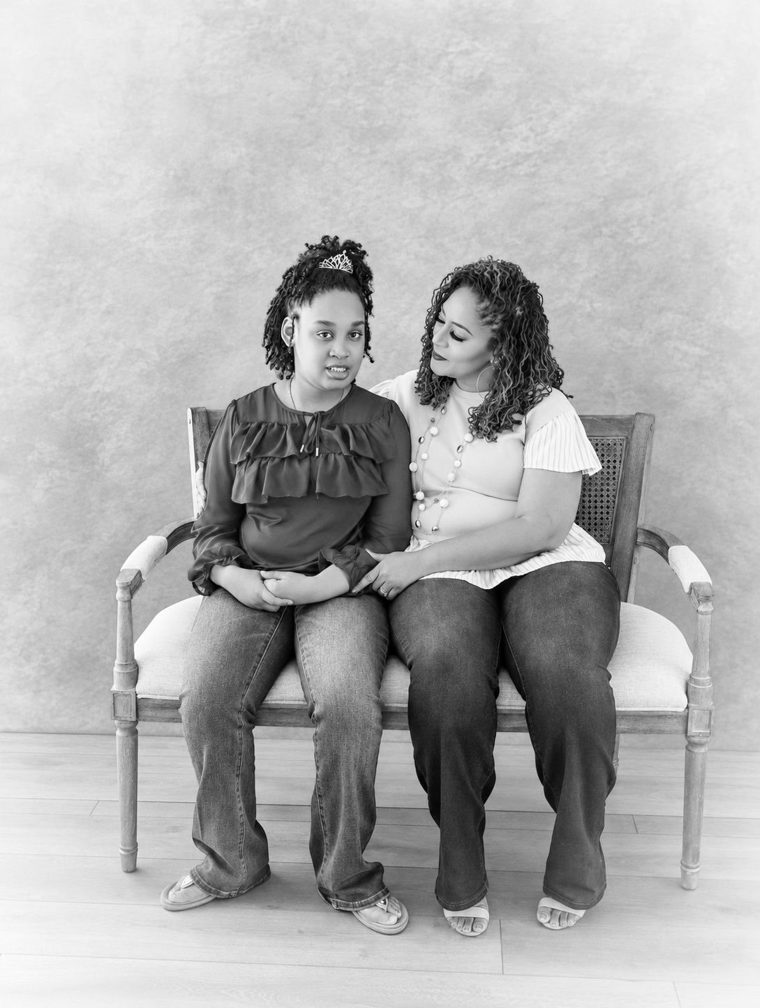 Black and white portrait of two women with curly hair sitting close together on a vintage wooden bench in a studio.