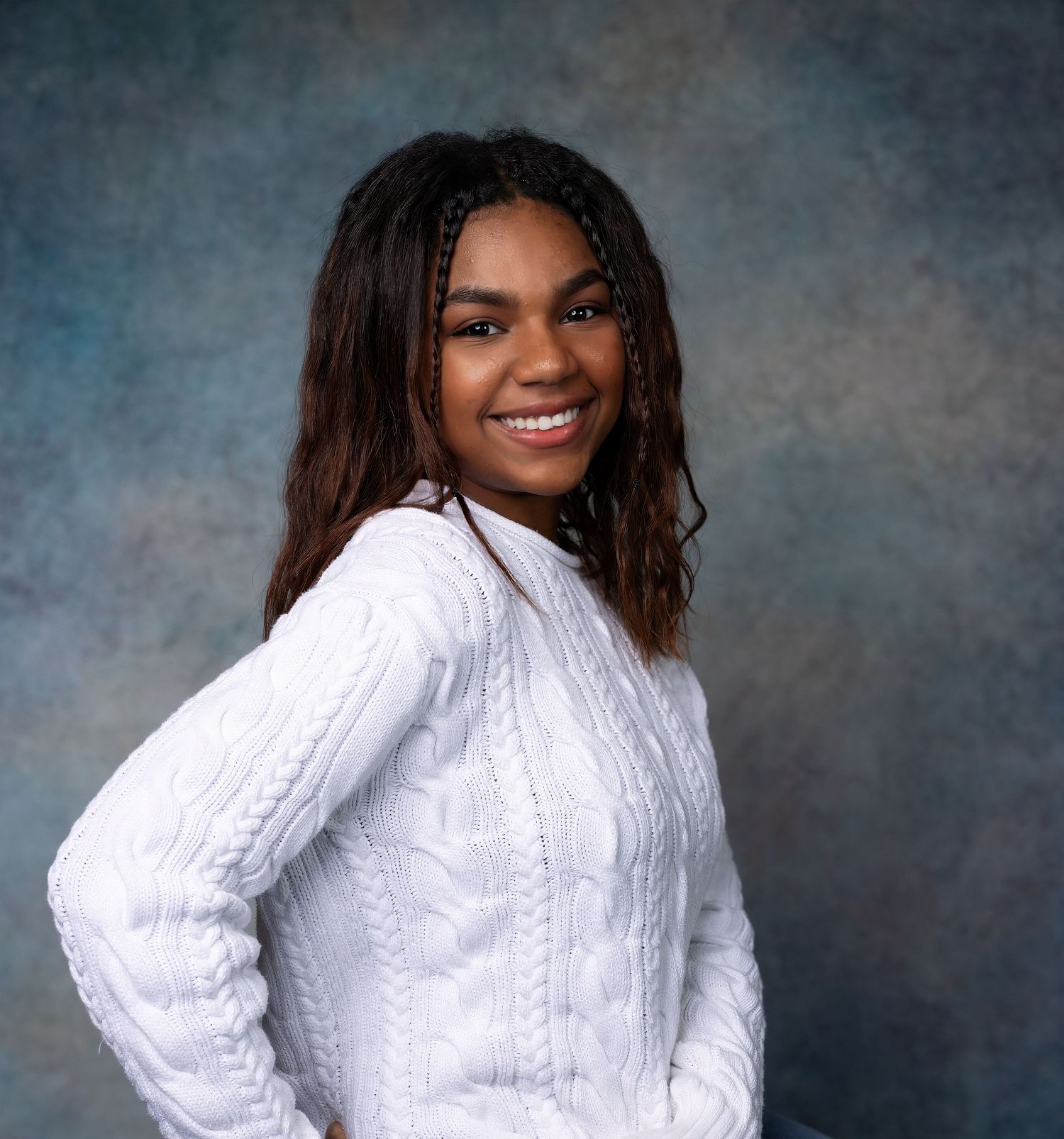 Professional portrait of someone in a white cable knit sweater against a blue-grey backdrop.