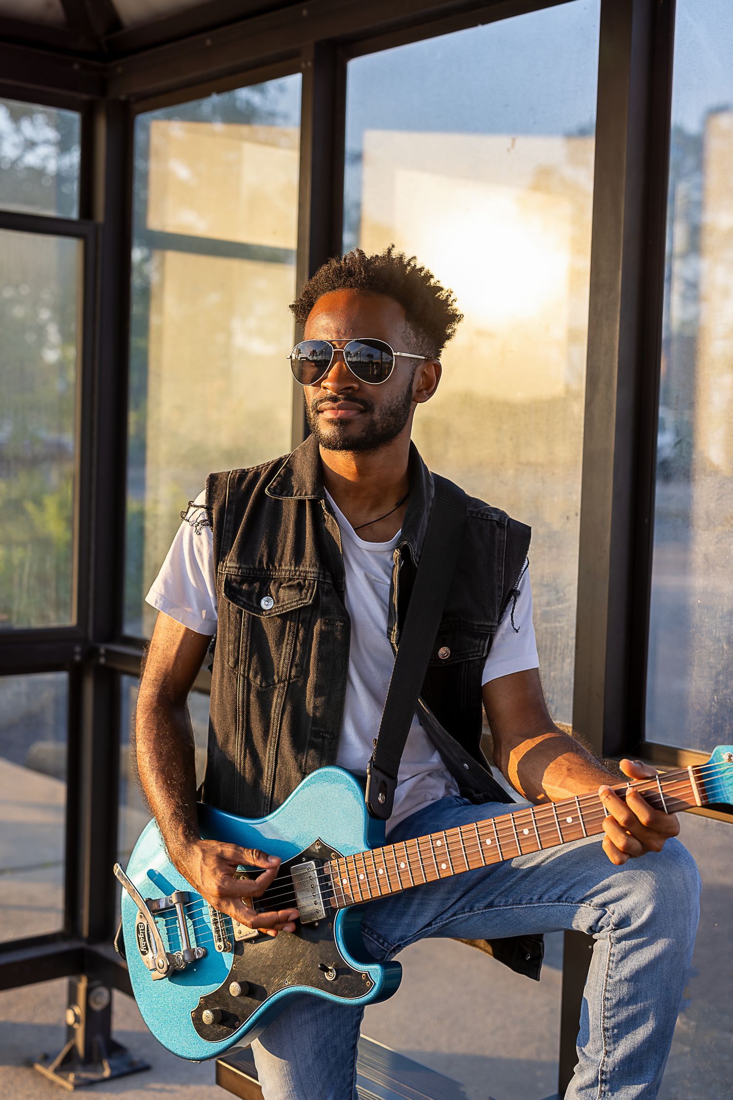 A musician plays a turquoise guitar while sitting in a sunlit glass enclosure.
