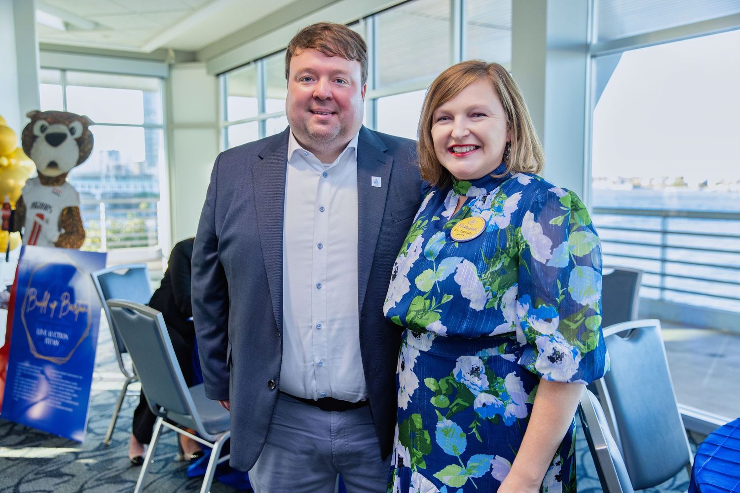 Two professionals stand together smiling in a bright office space with blue decor and windows in the background.