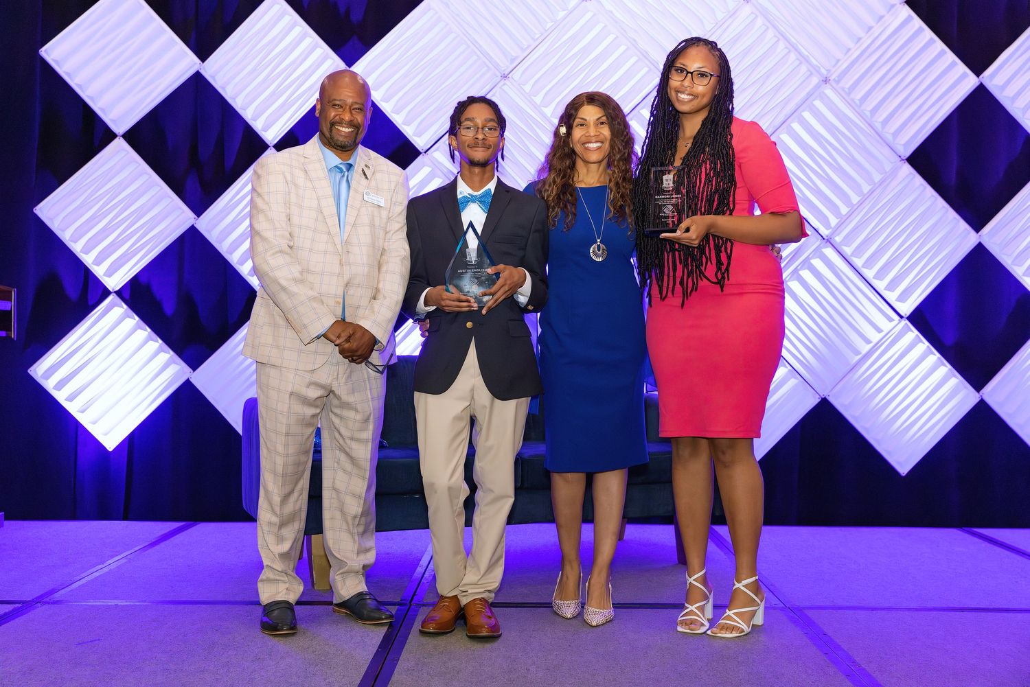 Four people pose together on stage at a formal awards ceremony, one holding a glass trophy award.