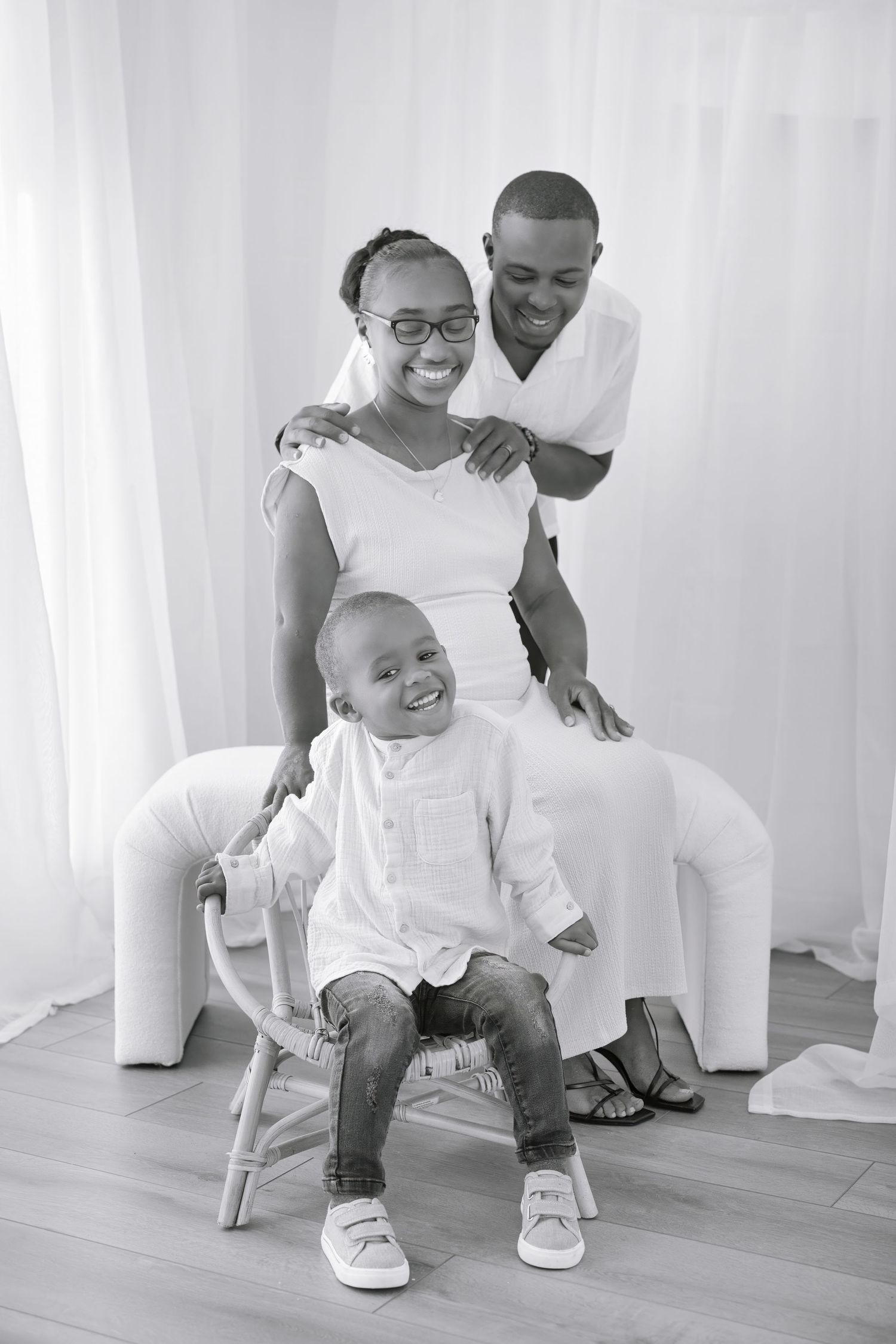A joyful family photo in black and white showing parents and a smiling toddler sitting on a chair against light curtains.