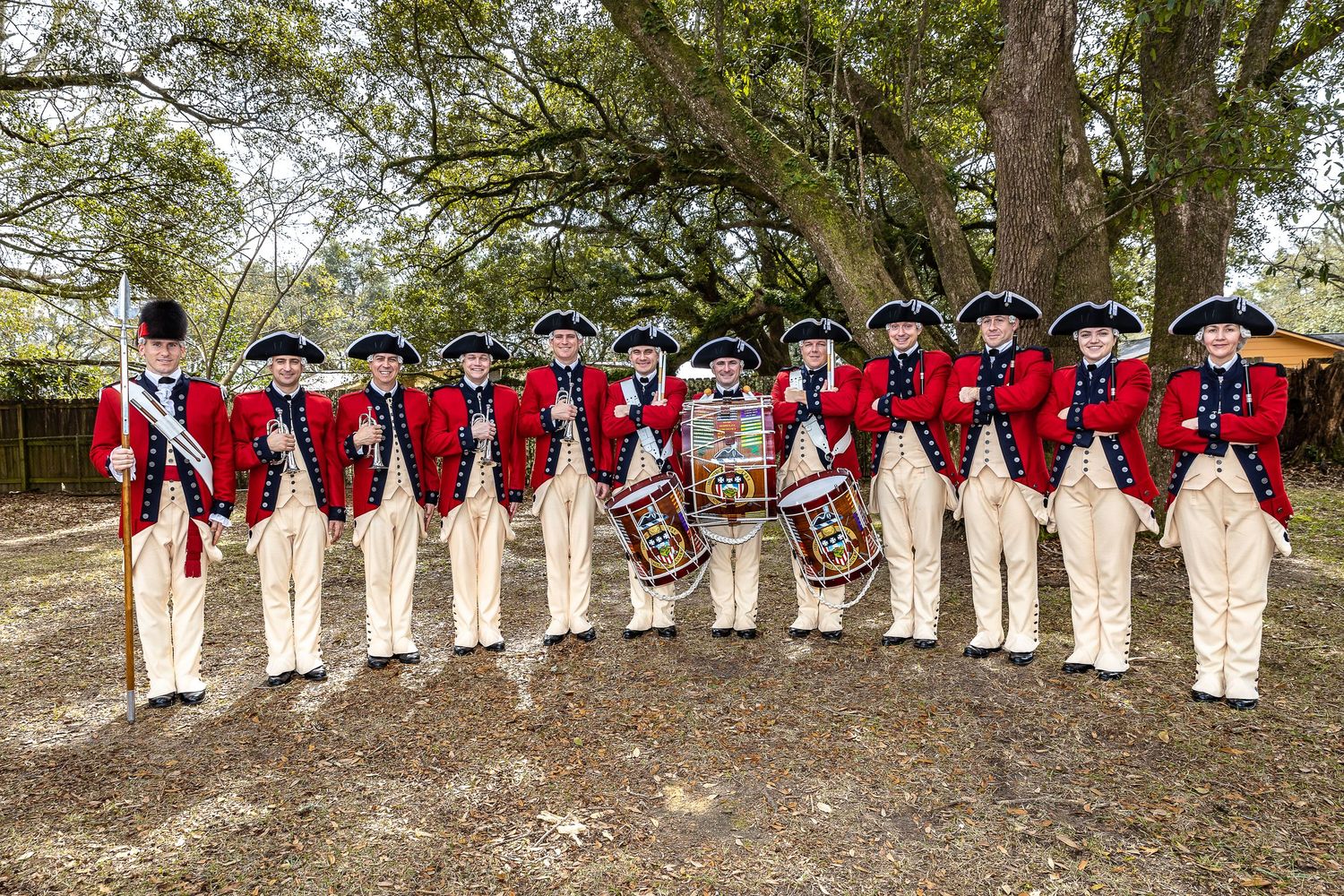 A line of historical reenactors in red colonial military uniforms pose outdoors under large oak trees.