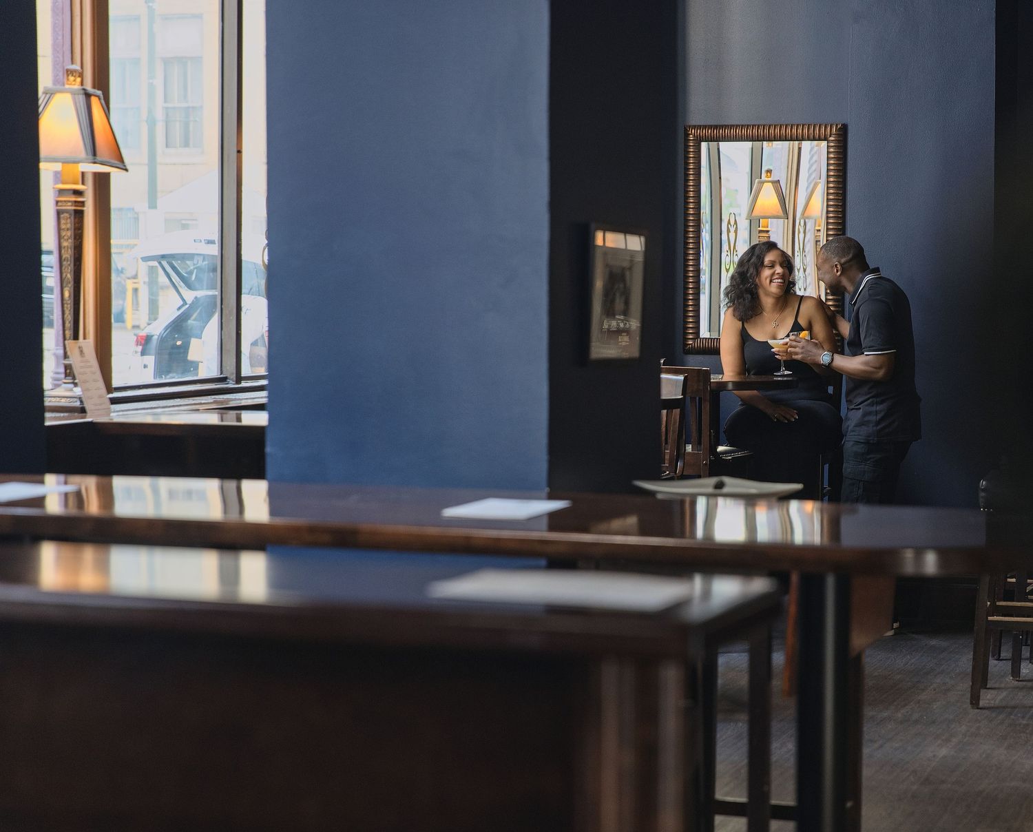 Couple sharing drinks at a cozy blue-walled bar restaurant near a window in intimate conversation.