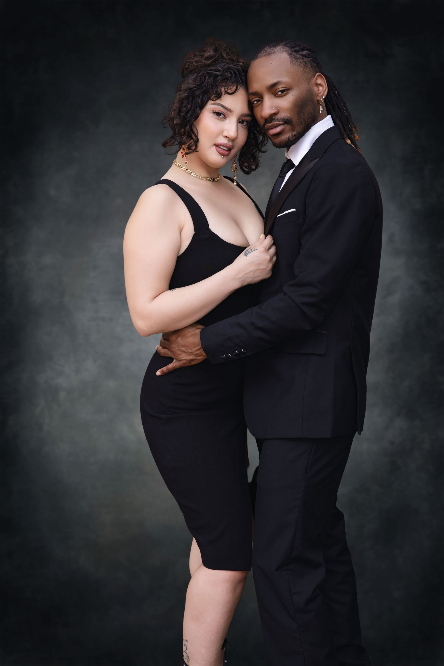 Romantic studio portrait of a couple in formal black attire against a dark grey background.