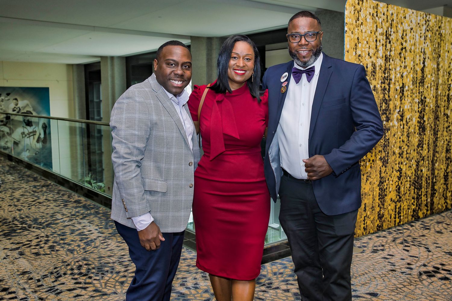 Three people in formal attire posing together at an event with gold backdrop.