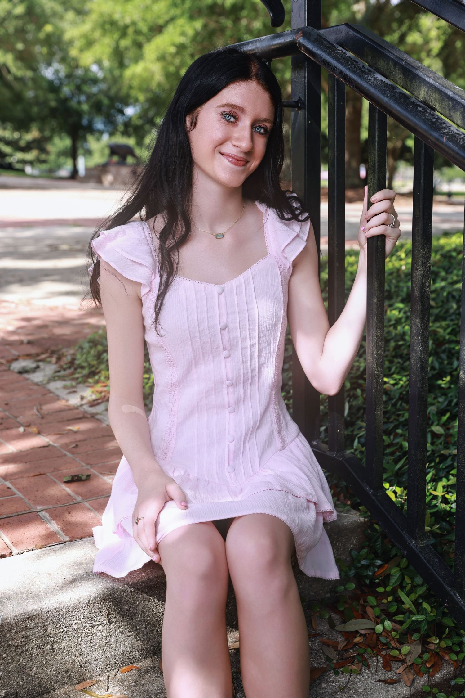 Someone in a light pink sundress sits on outdoor brick steps while holding onto a black metal railing in a garden setting.