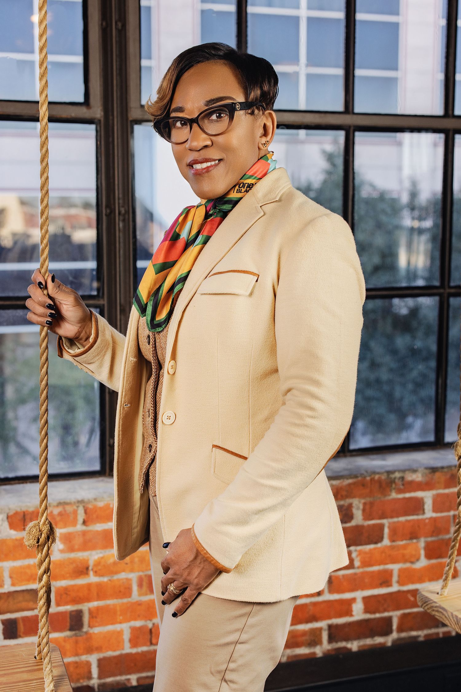 Professional wearing a cream blazer and glasses poses playfully on a rope swing against brick and window backdrop.