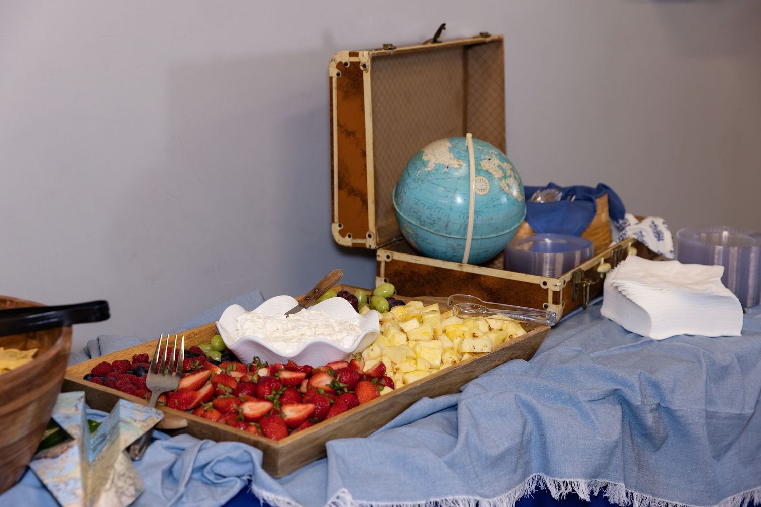 A travel-themed food display with fresh strawberries, fruit, and honey beside a vintage suitcase and globe centerpiece.