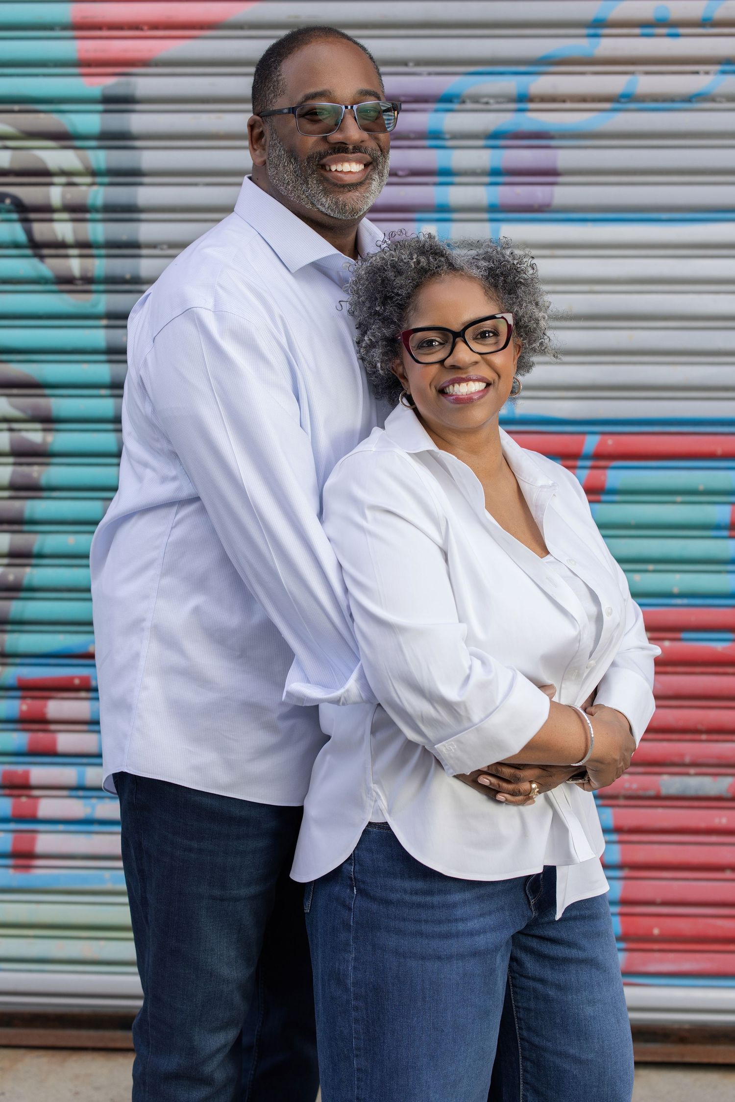 Smiling couple in white shirts and jeans posing together in front of colorful graffiti wall.