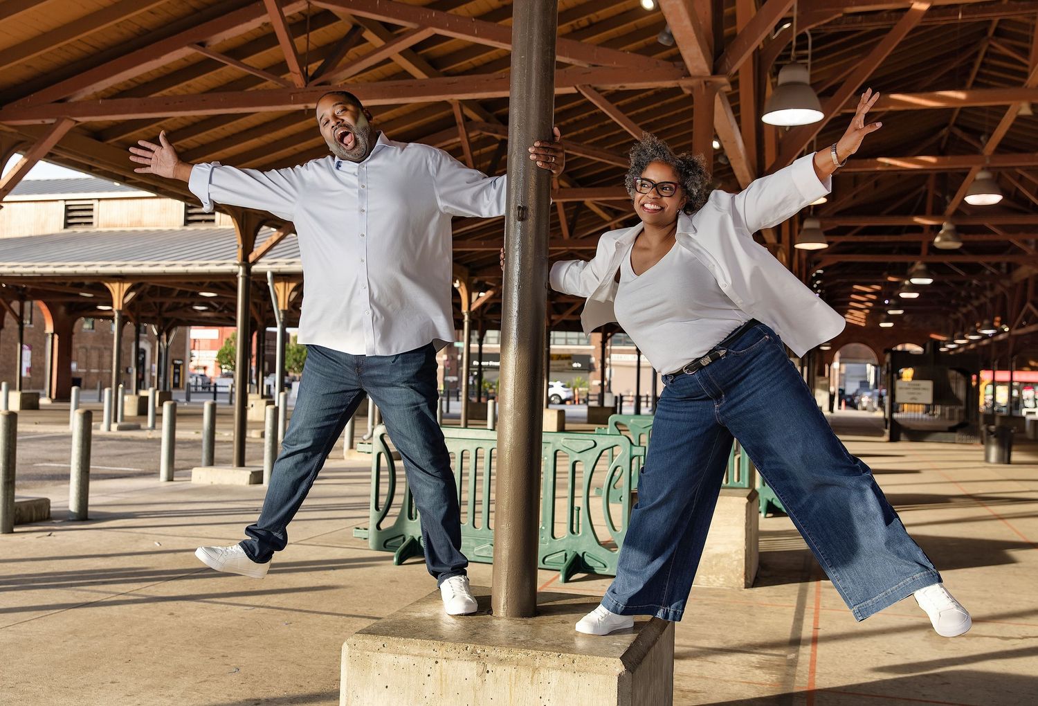 Two joyful people strike fun jumping poses under a wooden pavilion at an outdoor market, wearing casual white shirts and jeans.